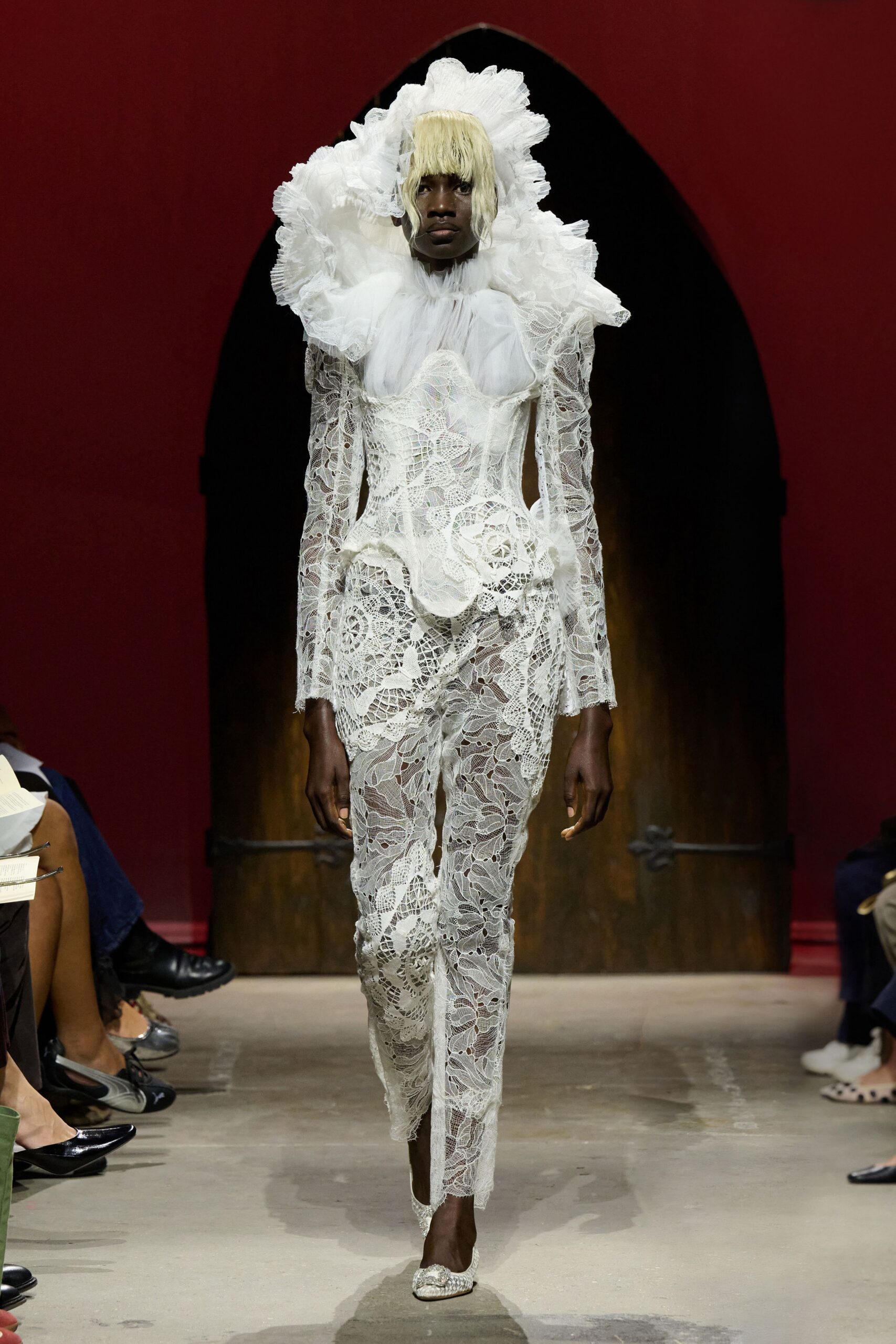 Monumental white organza ruff, edges knife-pleated into a cloud. Sheer white tulle bodice under corded lace with shell-like hip scrolls. Slim white lace trousers, one hem falling into a pointed ankle flap. White lace flats with a beaded buckle. Crossing a chapel aisle after the flowers have started to wilt.
