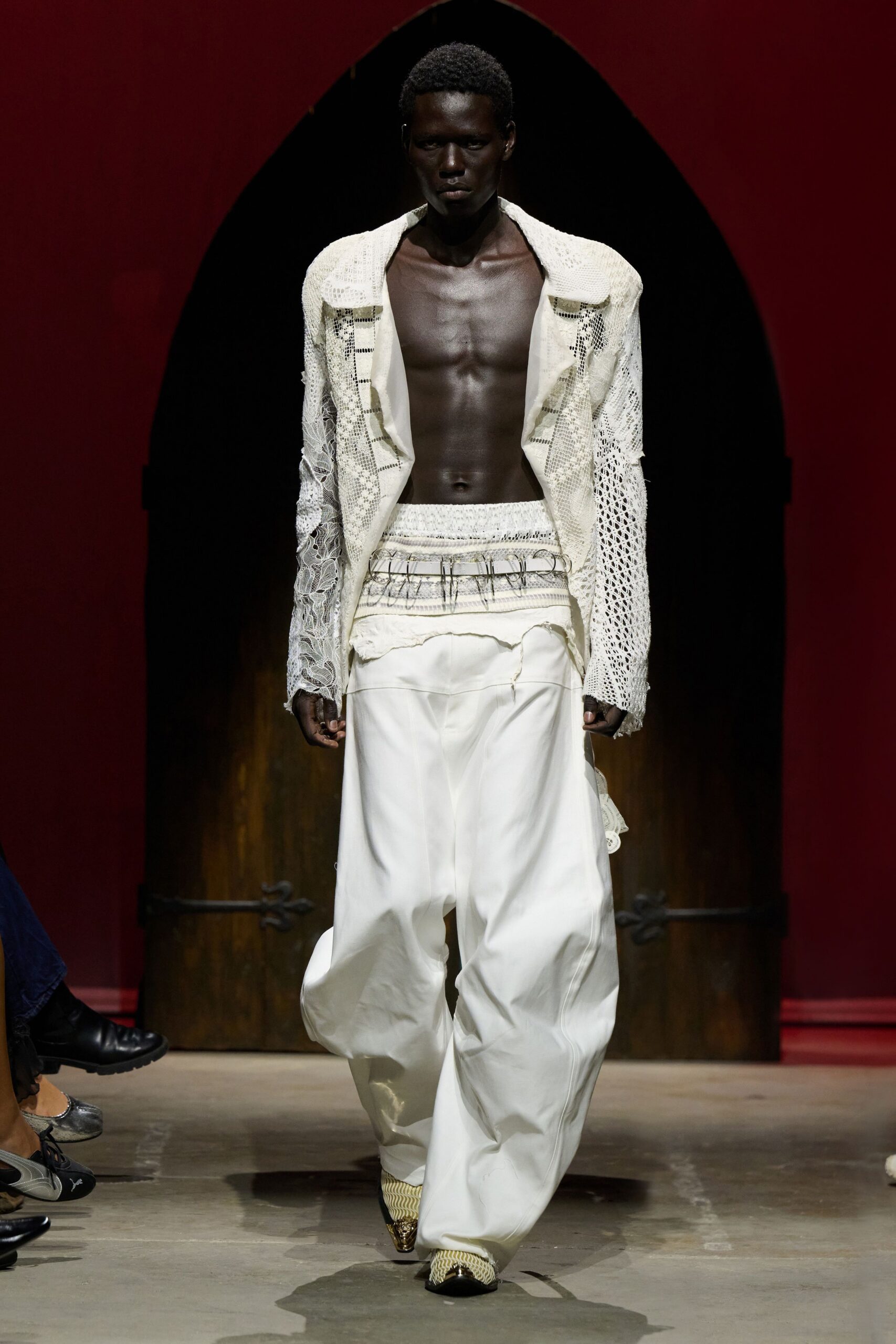 Open ivory crochet jacket, one sleeve in denser floral lace. Layered openwork bands ringed with bent silver wire loops. Billowing white trousers, raw flap hanging loose below the waist. Cream woven boots with pointed metal-capped toes. Crossing a monastery courtyard after rain, hems picking up the damp.