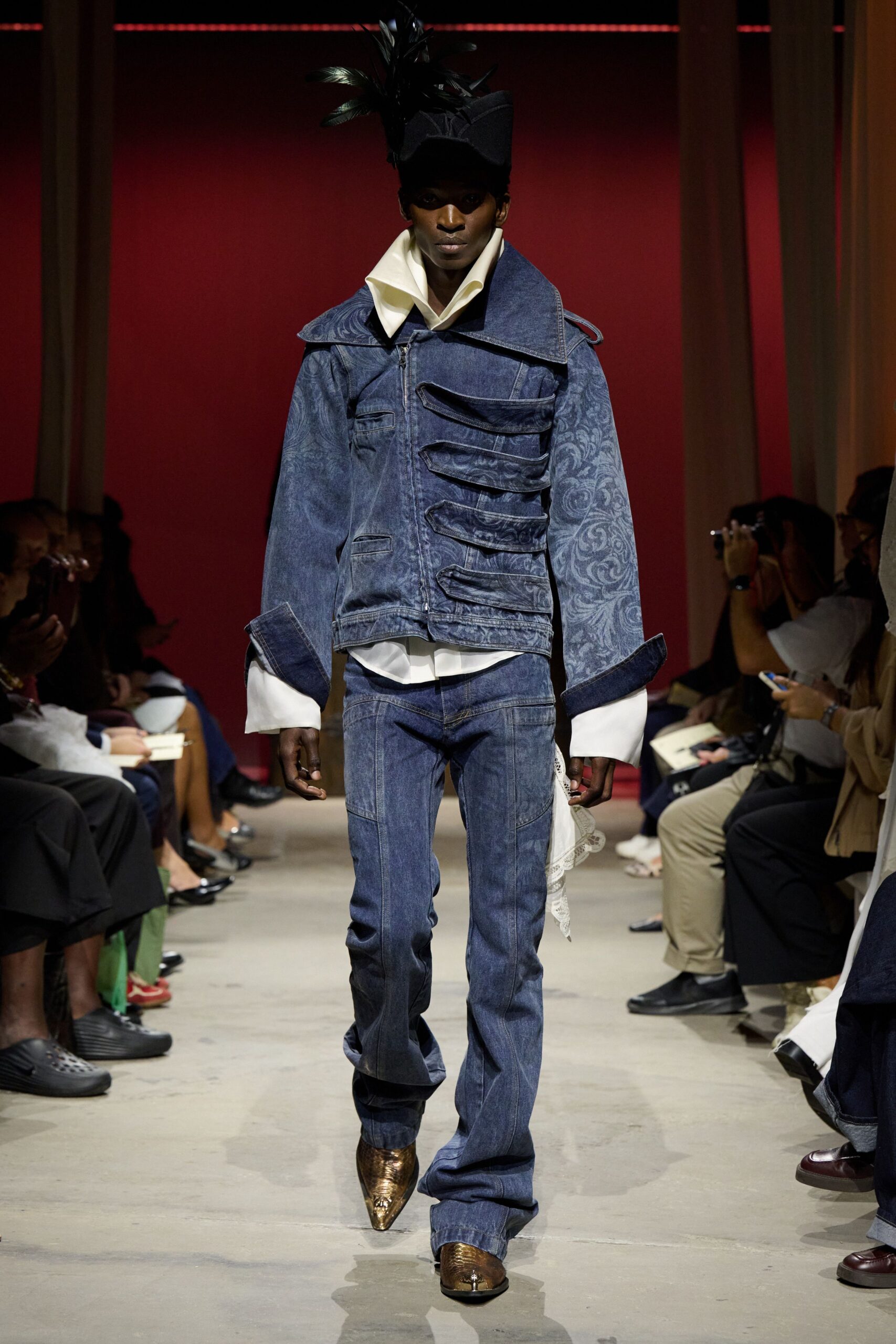 Black felt bicorne with a burst of upright feathers. White shirt, collar flared high and cuffs hanging long. Washed indigo denim jacket, one side stacked with looping flap bands. Matching bootcut jeans with curved panel seams. Pointed bronze snakeskin boots. Crossing a manor hallway after midnight, still in yesterday's clothes.