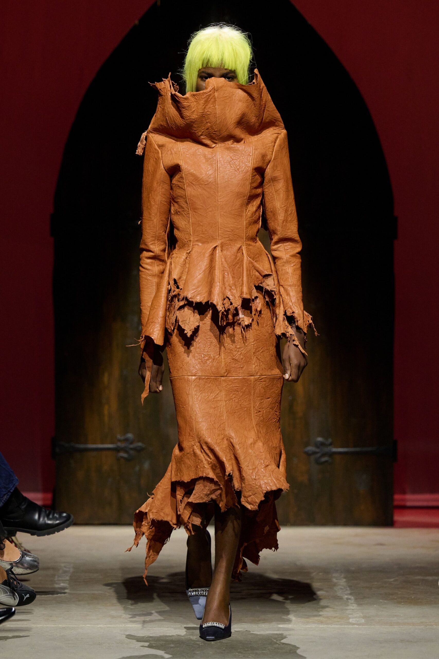 Acid-lime bob wig cut blunt across the brows. Cognac leather dress with a towering funnel collar and shredded peplum edges. Sheer black stockings. Square-toe pumps with crystal-trimmed vamps. Church basement pageant, someone left the prop dragon by the vestry door.