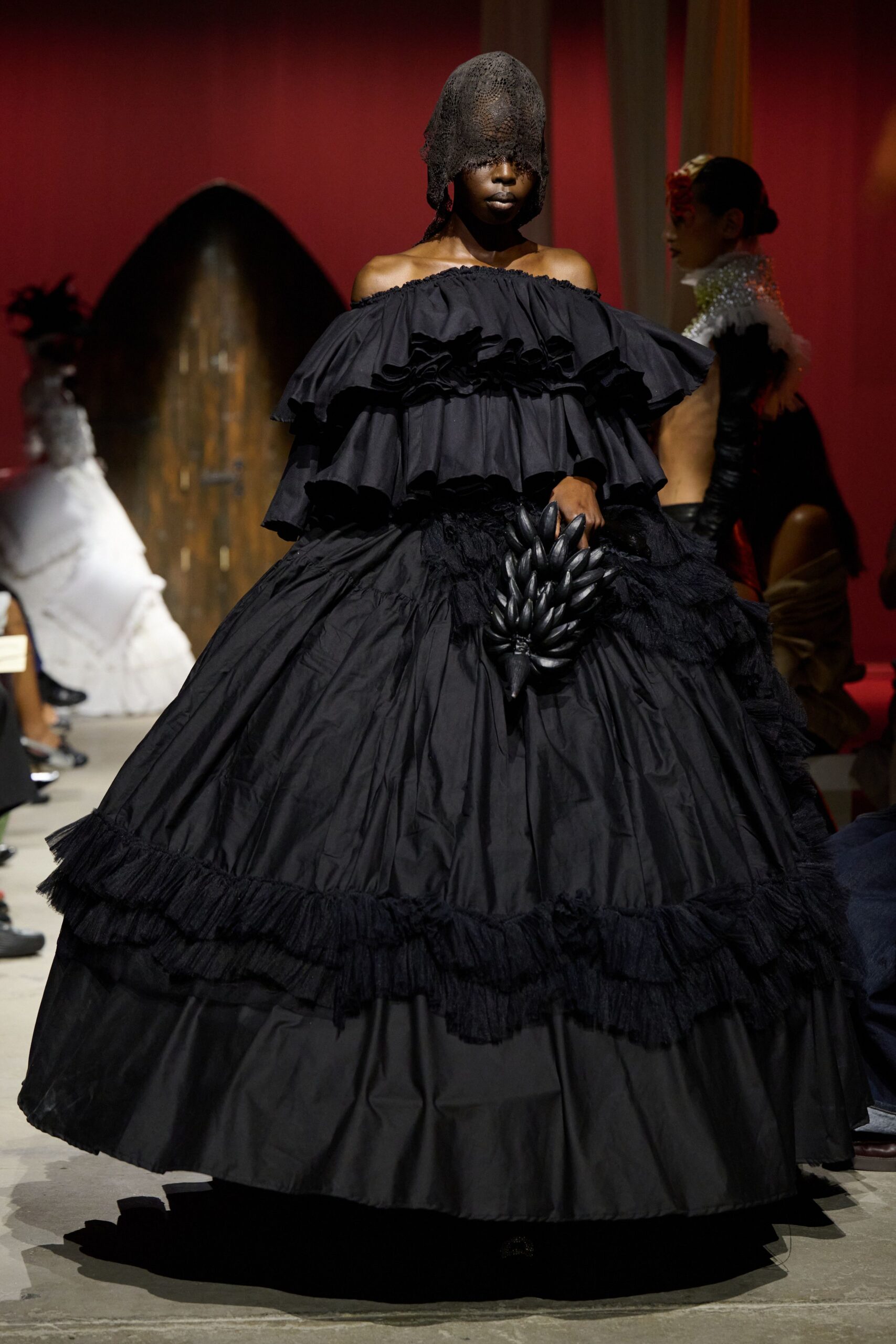 Openwork black veil dropped low over the eyes, hem ragged. Off-shoulder black gown with stacked flounces and a vast crinkled skirt. Dense frayed fringe circling the lower tiers. Black cluster bag shaped like a hanging bunch of bananas. At a candlelit banquet where the fruit centerpiece keeps going missing.