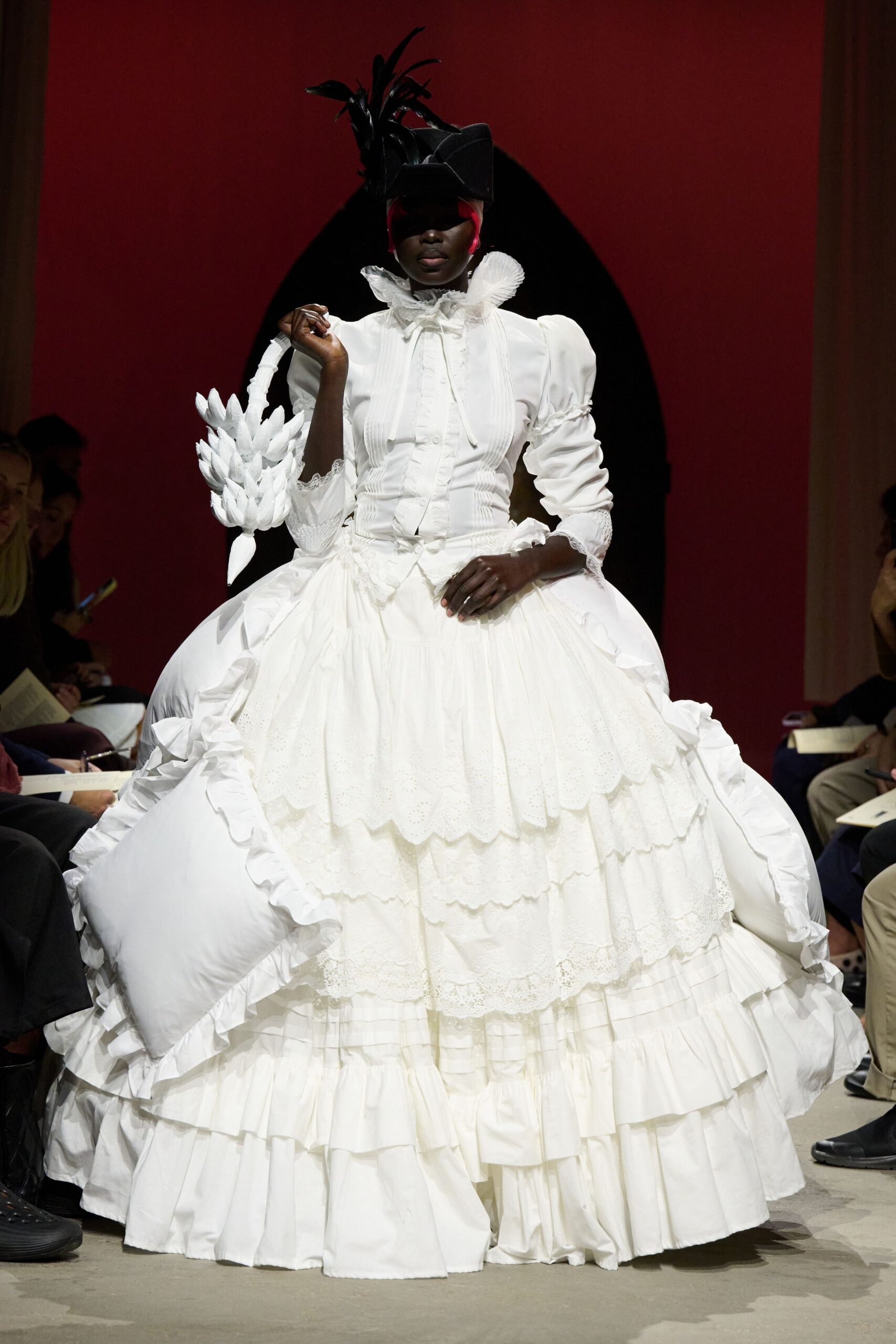 Black folded hat with a high spray of lacquered feathers. White fitted blouse, pintucked front and pleated ruff framing the face. Vast white skirt in scalloped broderie tiers, pillow-like panniers ruched at the sides. White petal-cluster handbag on a wrapped handle. For arriving at a masked ball through the kitchen garden.