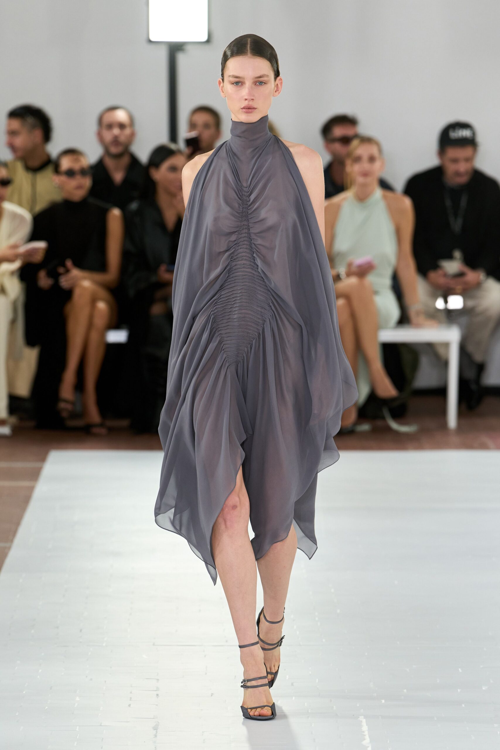 Smoke-gray chiffon halter dress, ruched down the center into a pointed drape. Charcoal strappy sandals with a tiny silver buckle at the ankle. Crossing a museum atrium while the air-conditioning finally kicks in.