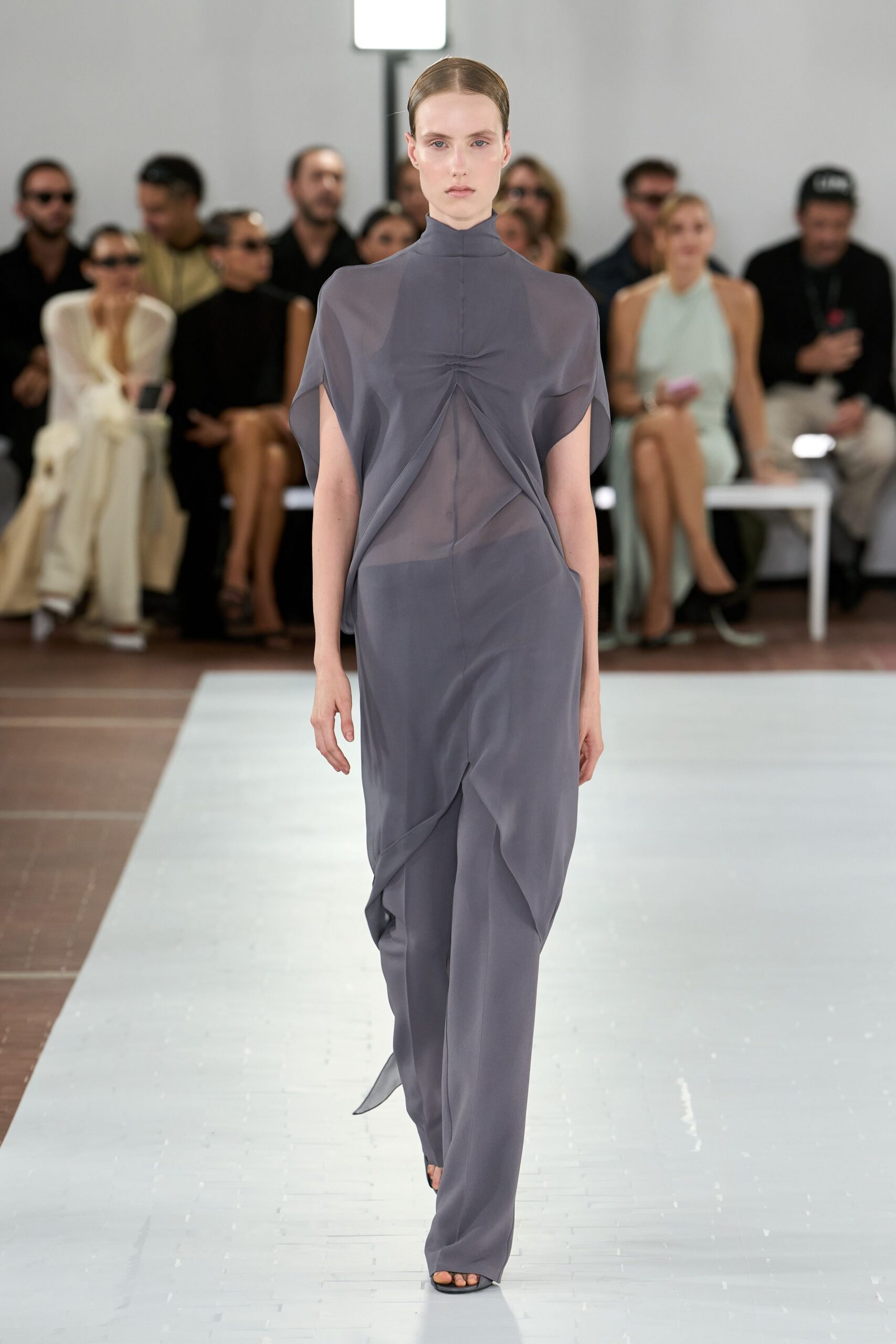 Smoke-grey sheer jersey tunic, pinched at the sternum into a soft twist. Matching fluid trousers disappearing under the tunic's angled split hem. Black barely-there sandals. Crossing a museum atrium before the air-conditioning has caught up.