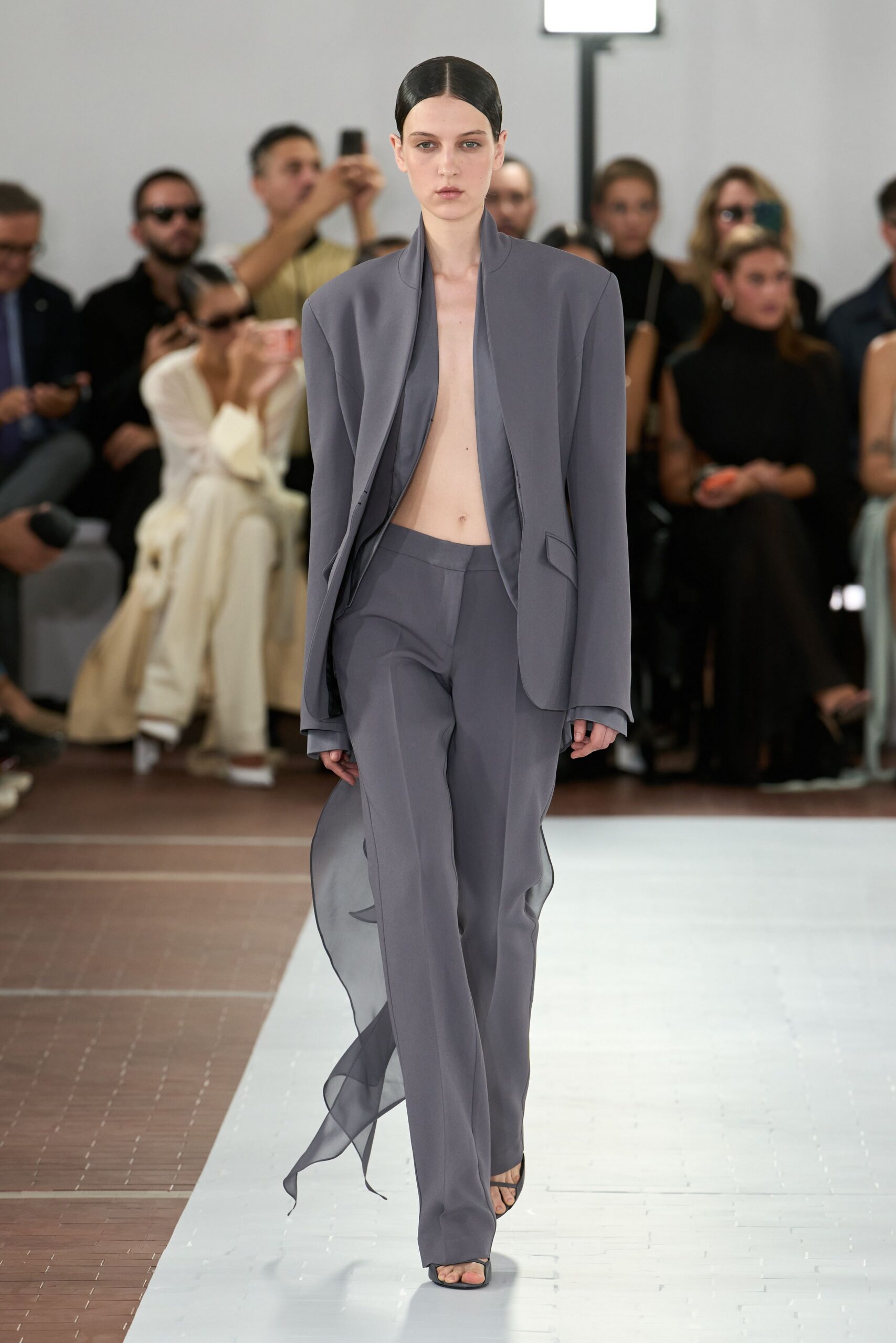 Slate crepe jacket cut open to the navel, with a curved standing collar. Matching straight trousers with sheer chiffon godets trailing from the side seams. Black thong sandals with a square toe. Crossing a museum atrium before the air conditioning fully wakes up.