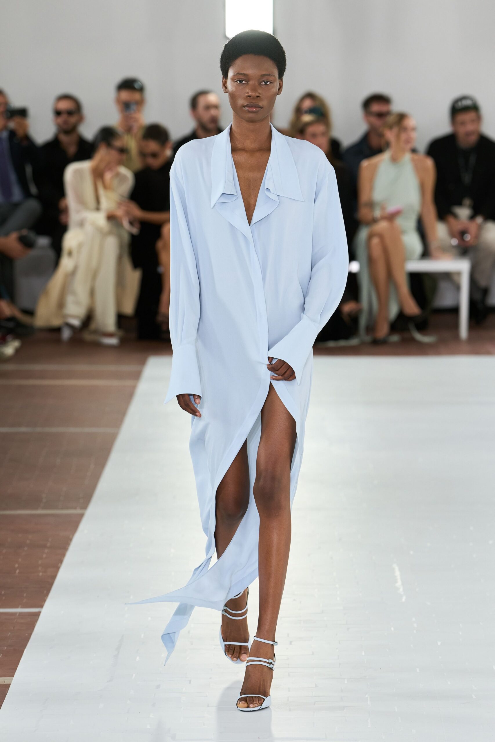 Powder-blue crepe dress with a deep draped lapel and trailing hem. White barely-there straps, one buckle glinting at the ankle. Crossing a hotel terrace before breakfast, pool tiles still cold.