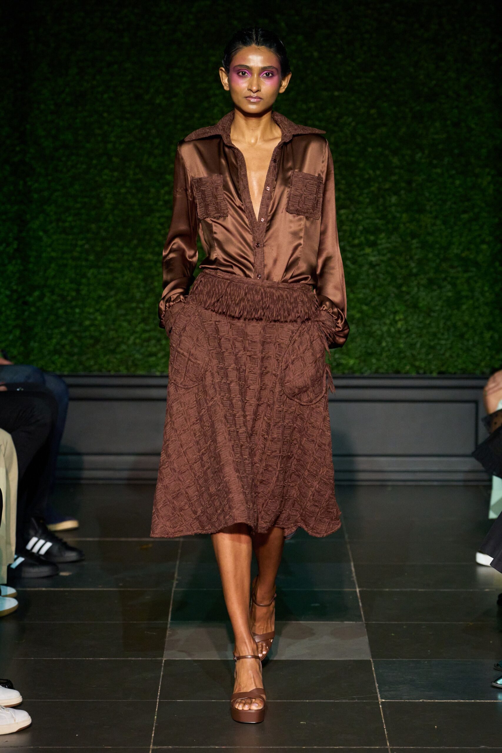 Chocolate satin blouse with basket-weave trim edging the deep placket. Matching crinkled weave midi, fringed waistband slanting over one hip pocket. Brown platform sandals with slim ankle straps. For lunch on a shaded terrace after the rain has steamed off.