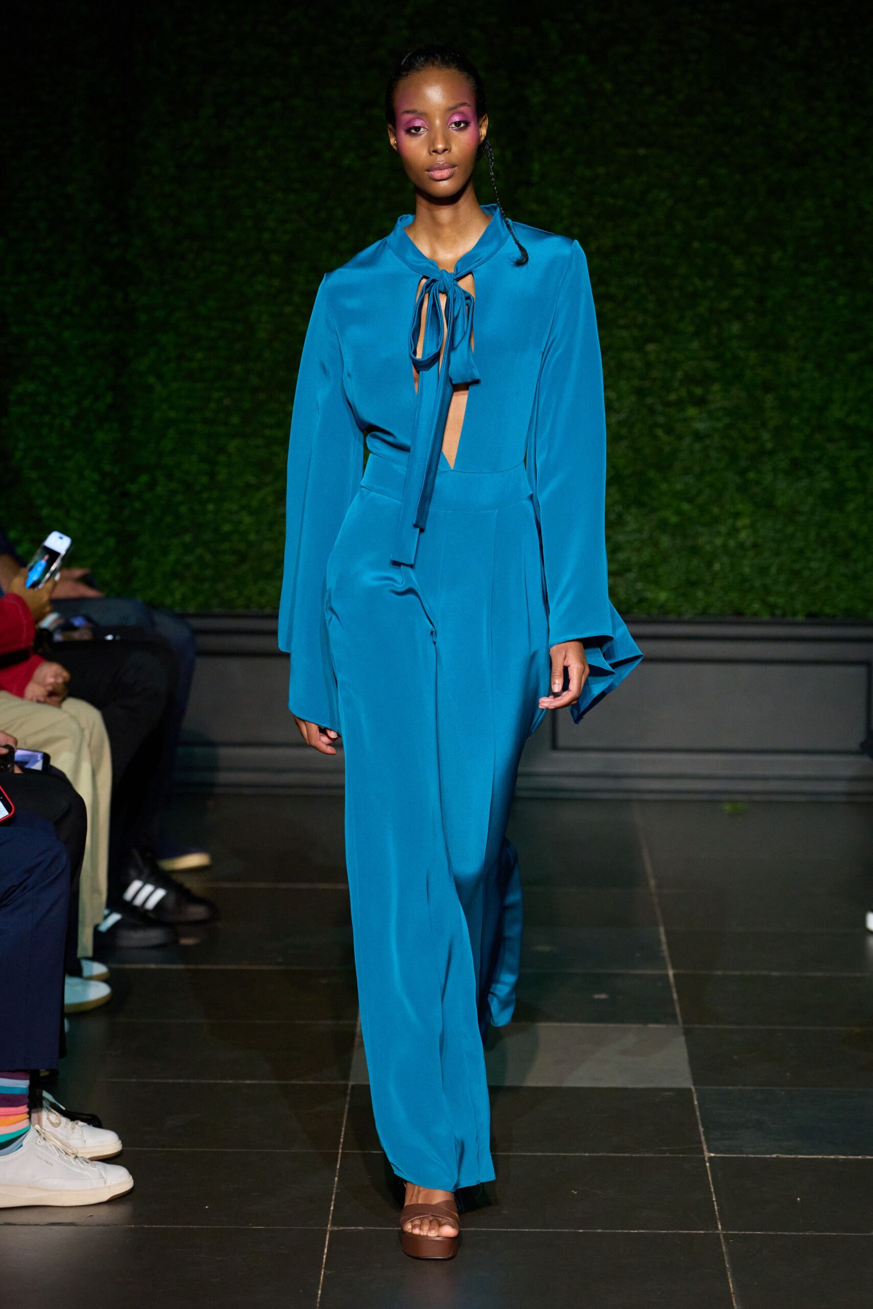 Peacock-blue crepe jumpsuit with a slit bodice and loose neck ties. Long flared sleeves dropping past the wrist in a soft drape. Brown platform sandals with a broad strap across the toes. For crossing a tiled courtyard just after the hotel bar opened.