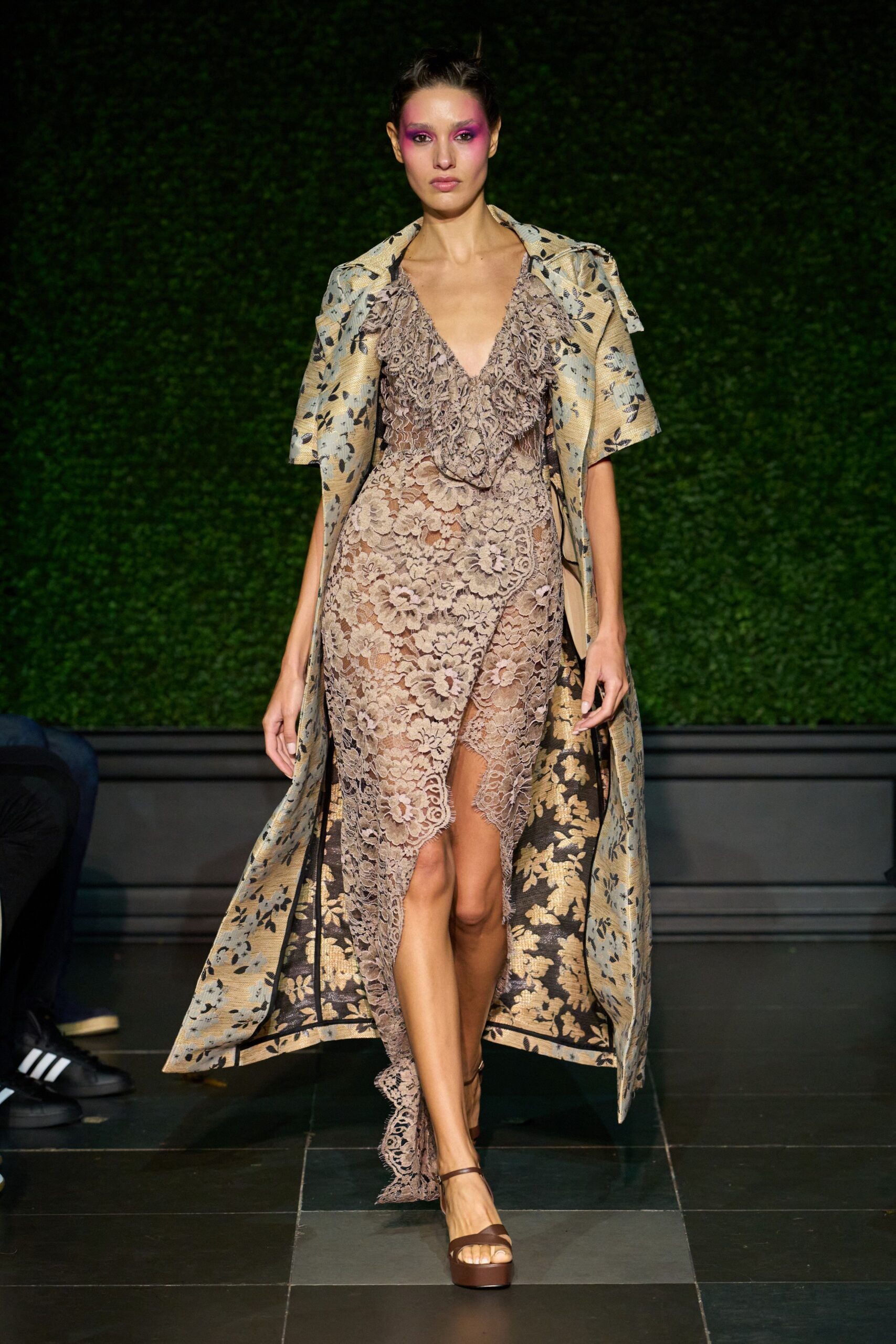 Long gold brocade coat with black floral lining flashing at the split front. Taupe floral lace slip dress, ruffled V neckline and uneven scalloped hem. Brown platform sandals with slim ankle straps. Garden wedding after midnight, grass damp and somebody still carrying champagne.