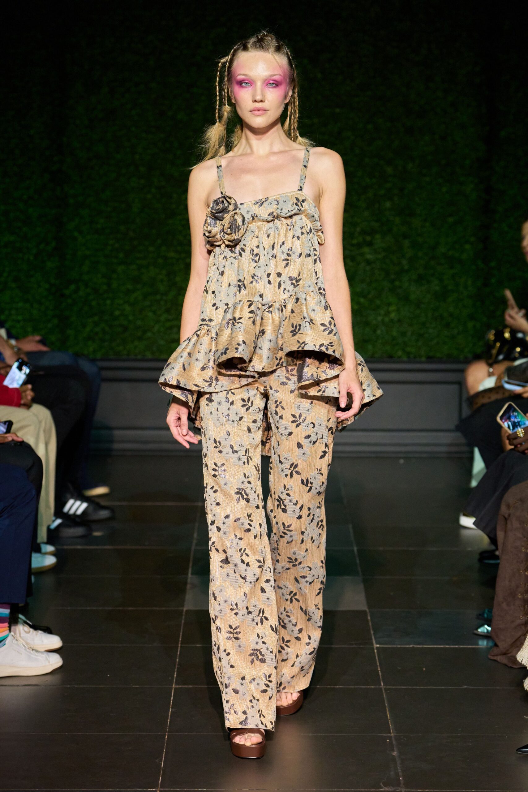 Beige crinkled floral top with rosettes knotted along the square neckline. Tiered flounce peplum jutting out in stiff ripples around the hips. Matching straight trousers in the same black leaf print jacquard. Brown platform sandals barely visible under the hem. Garden wedding spillover, walking back inside after dusk.