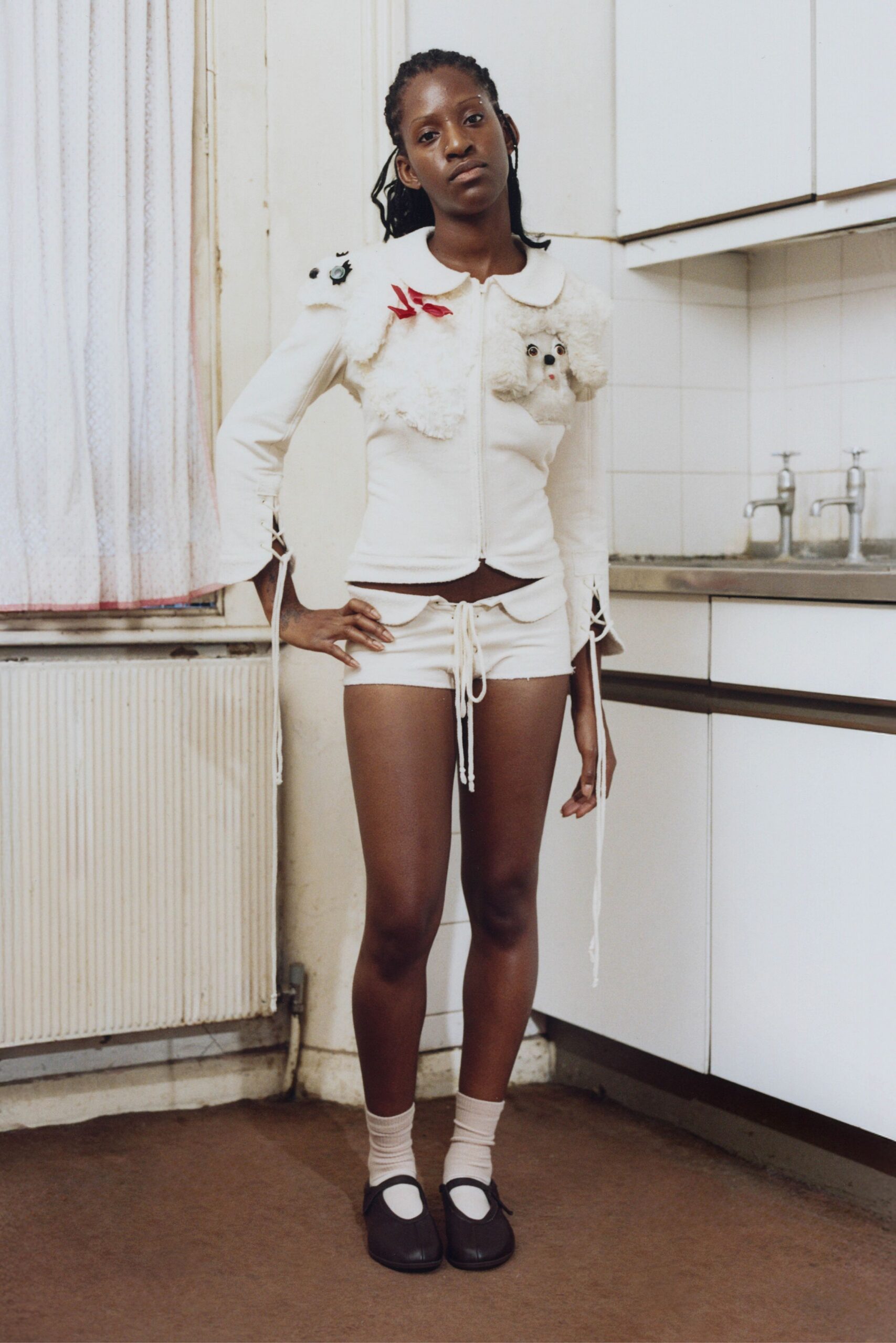 Cream zip cardigan with a Peter Pan collar and plush toy-dog appliques. Low-rise cream shorts, scalloped waistband tied with long drawstrings. Pale ankle socks scrunched softly above the shoe. Black square-toe Mary Janes with a narrow strap. In a rented kitchen after midnight, waiting for the kettle to click.
