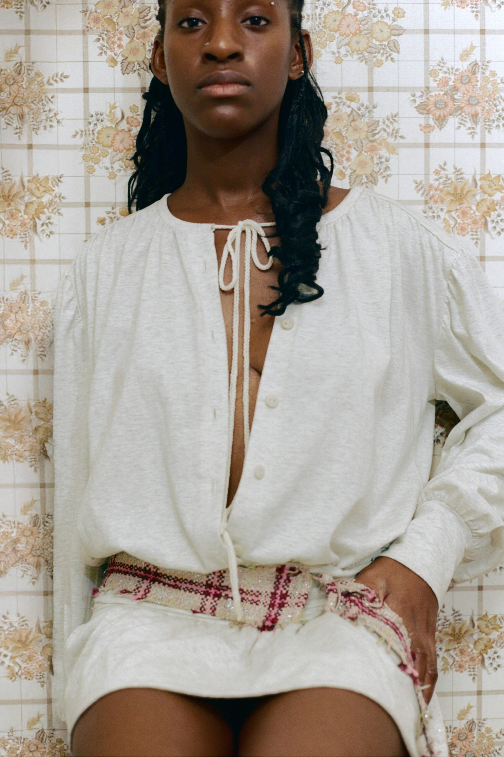 Cream slub-knit blouse with a cord-tied keyhole and low button placket. Matching cream shorts in the same soft heathered knit. Frayed pink-and-cream tweed sash, one ring glinting at the hip. Sunday afternoon at a rented cottage, wallpaper faded by three decades of sun.