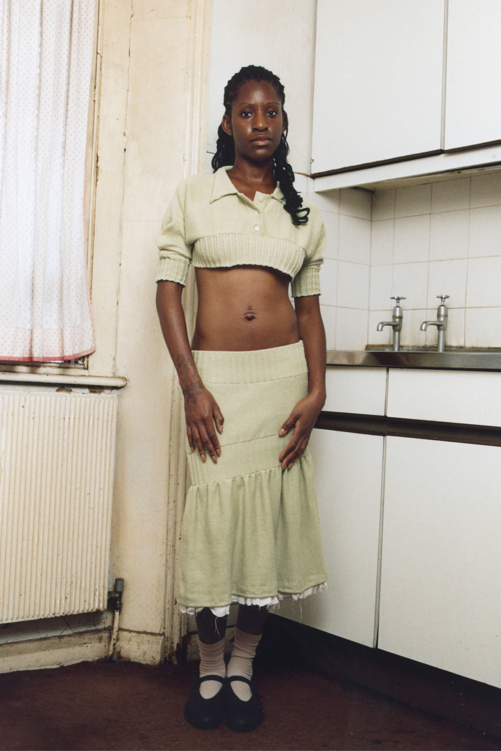 Pale pistachio knit polo, deep ribbing cinching the hem under the bust. Matching ribbed midi skirt, pieced tiers ending in a white scrap peek. Beige ankle socks slouched soft at the shin. Black Mary Janes with blunt rounded toes. Waiting for the kettle in a rented flat with someone else's curtains.
