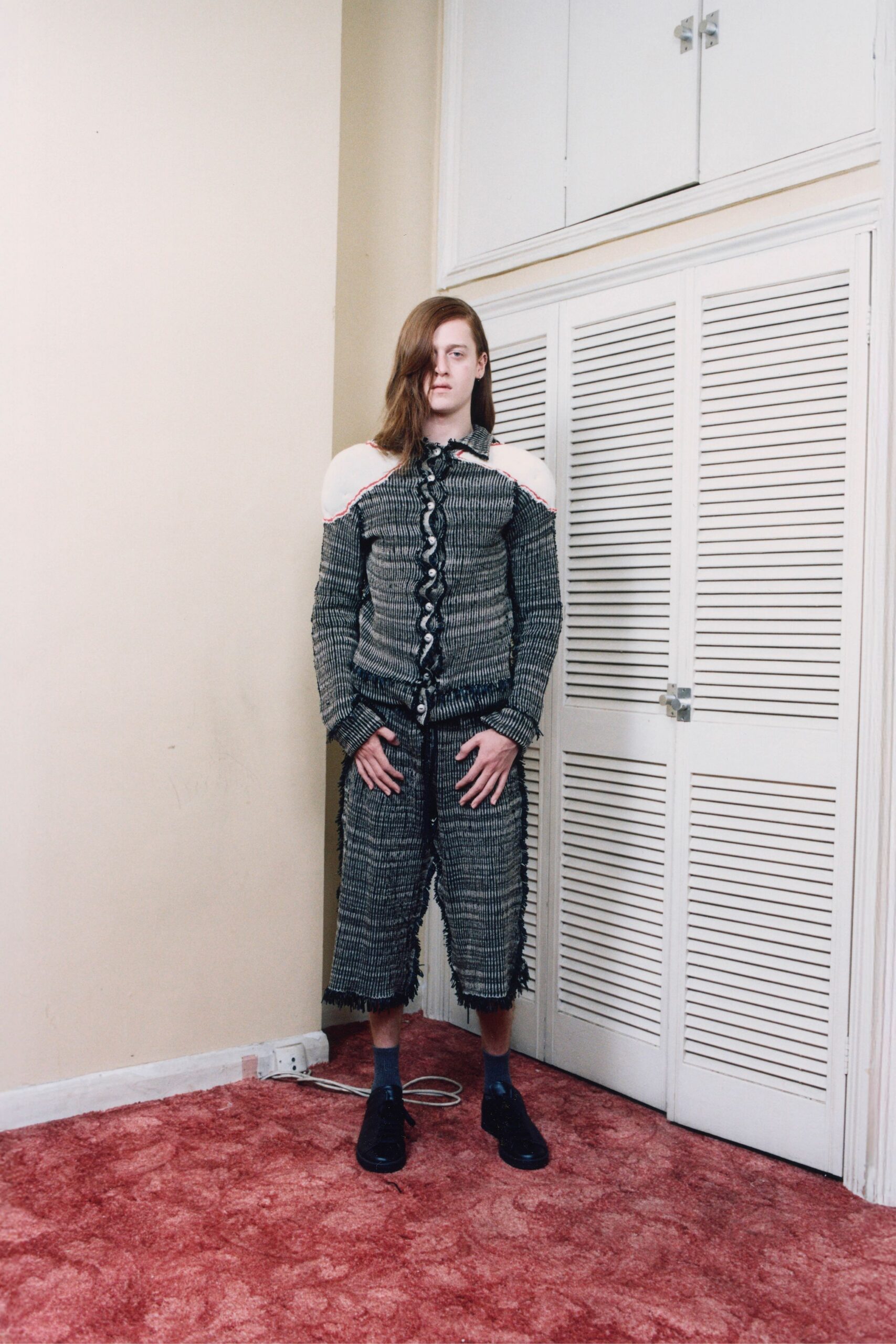 Grey puckered knit shirt, shoulders cut away and black ruffles zigzagging the placket. Matching checked cropped trousers, fringed seams running down both legs. Slate ankle socks. Black lace-up shoes with thick crepe soles. Standing in a rented room while the extension cord snakes across the carpet.