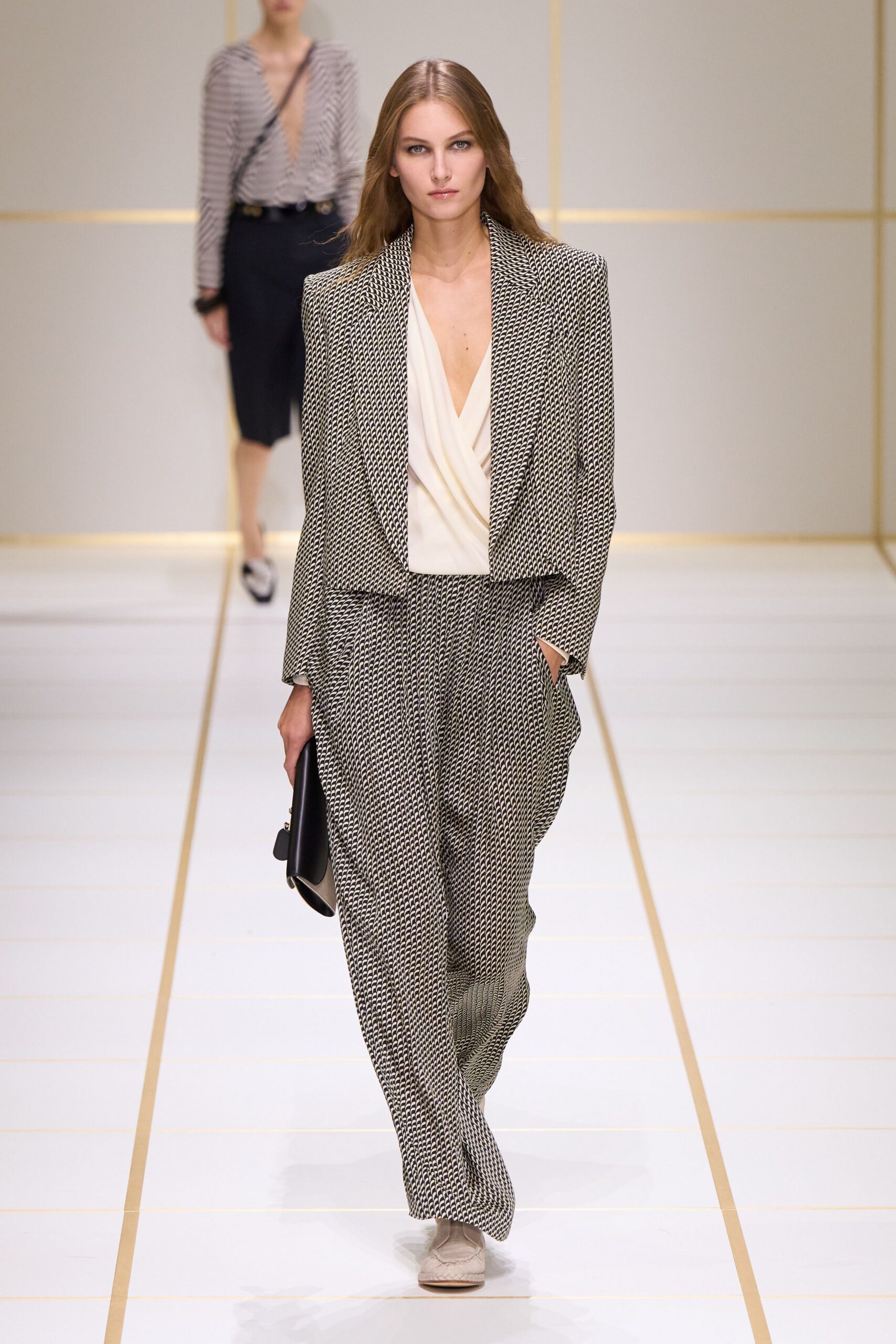 Boxy cropped jacket in black-and-cream chain-link jacquard. Ivory wrap blouse with a deep draped V neckline. Loose full-length trousers in the same chain-link weave. Flat black clutch with a rounded flap edge. Pale suede loafers with a moc-stitched apron. Walking into a hotel lobby before the conference coffee urns are empty.