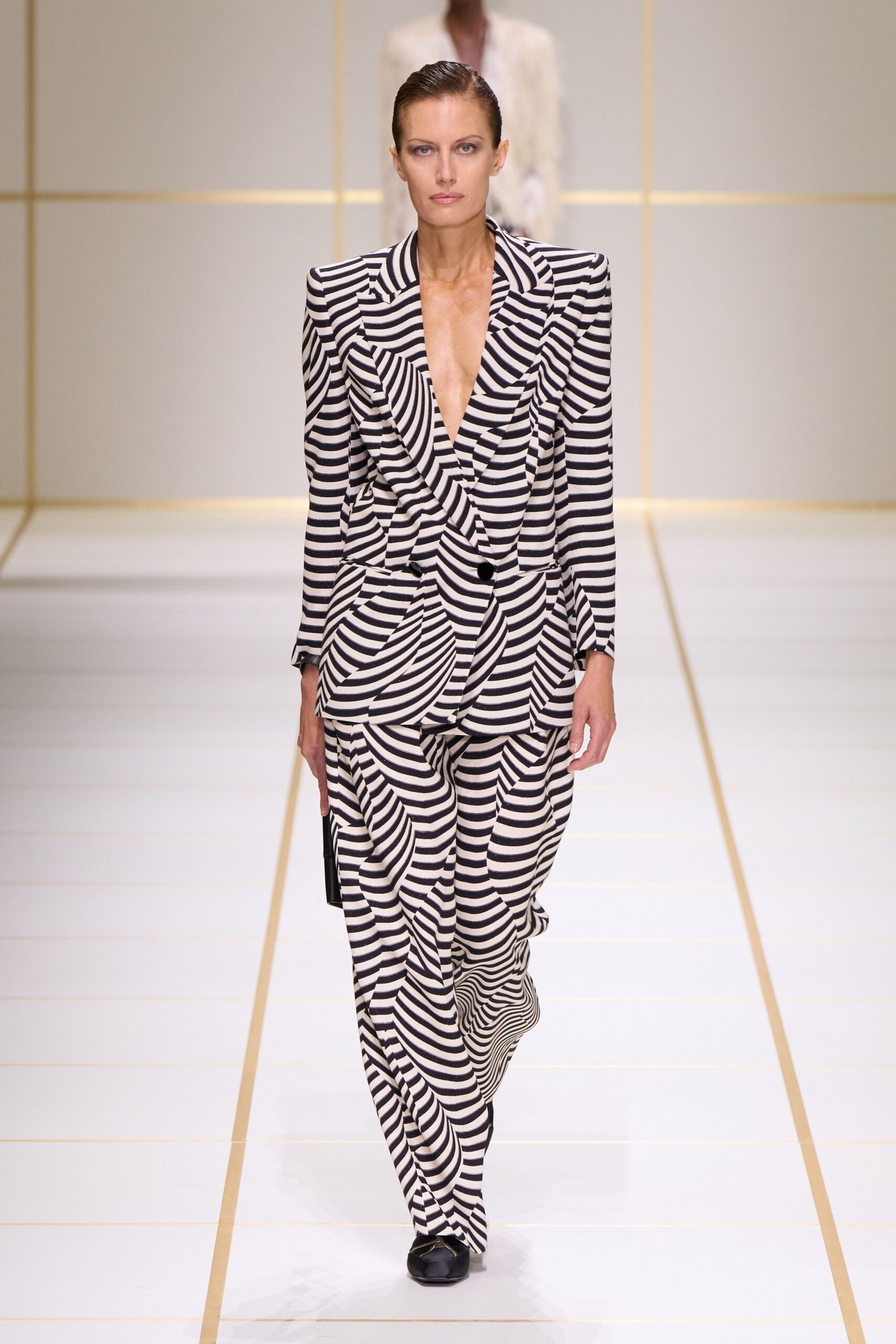 Black-and-ivory striped blazer, padded shoulders and plunging shawl lapels. Matching wide trousers, the warped stripes bending around each stride. Slim black clutch tucked straight at the side. Black square-toe flats with a pale cap catching light. Walking the hotel corridor carpet before anyone else is up.