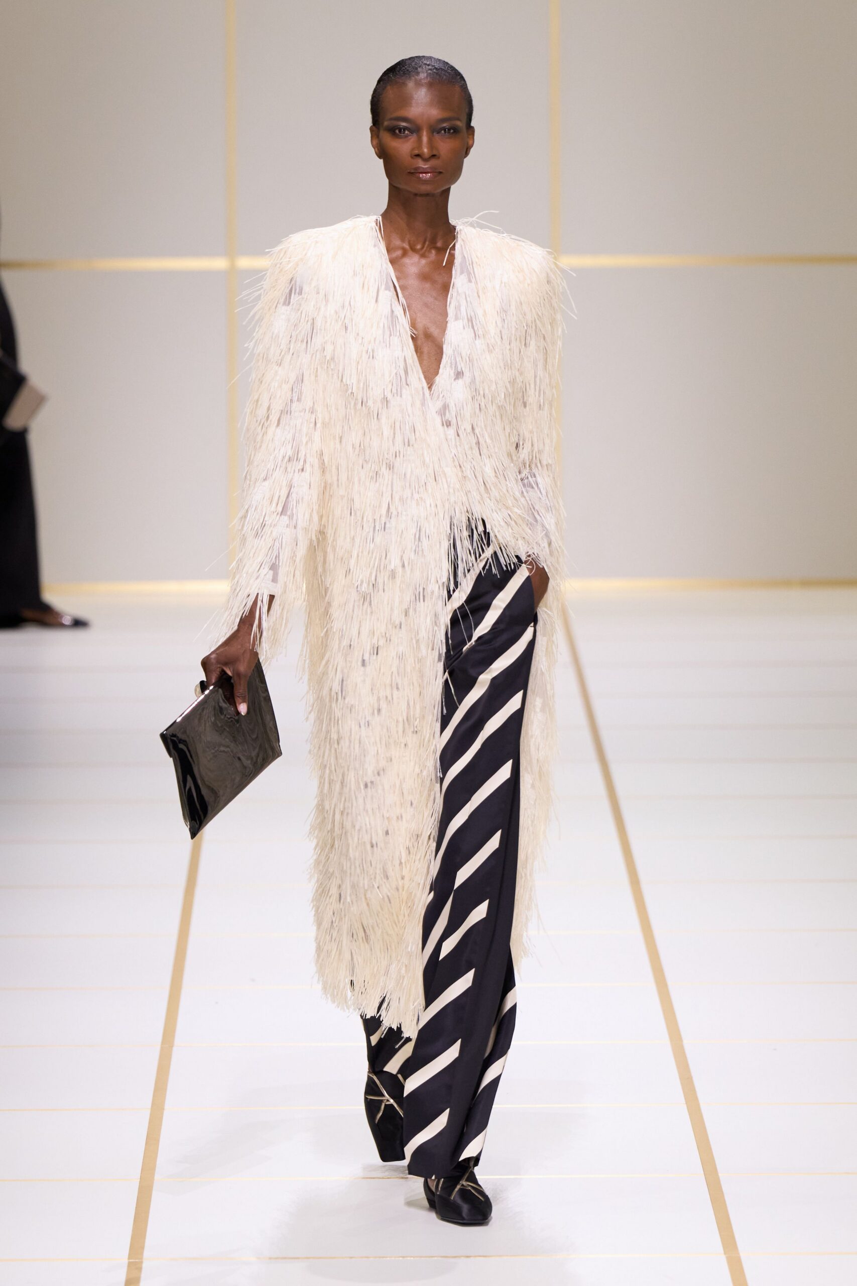 Ivory coat drowned in long raffia fringe, plunging open at the chest. Black wide-leg trousers slashed with broad ivory diagonal bands. Glossy black patent envelope clutch. Black square-toe flats with pale crisscross ties at the vamp. Crossing a hotel atrium after midnight, carrying the room key and nothing else.