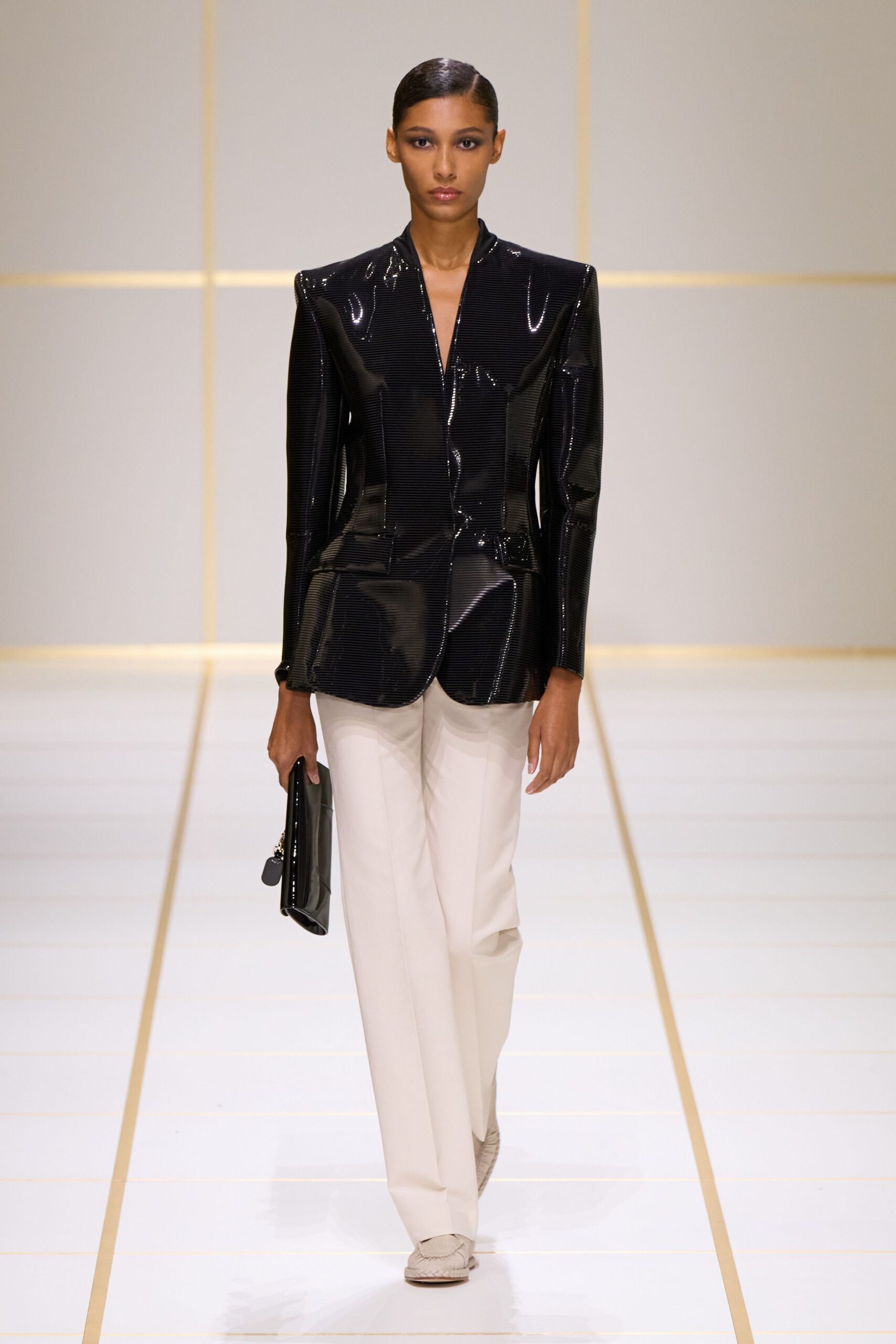 Black pinstriped patent jacket, shoulders squared hard and hem softly rounded. Cream straight-leg trousers with a pressed crease falling clean to the shoe. Glossy black foldover clutch with a small silver chain peeking out. White woven leather loafers. For crossing a hotel lobby before the breakfast tables are cleared.