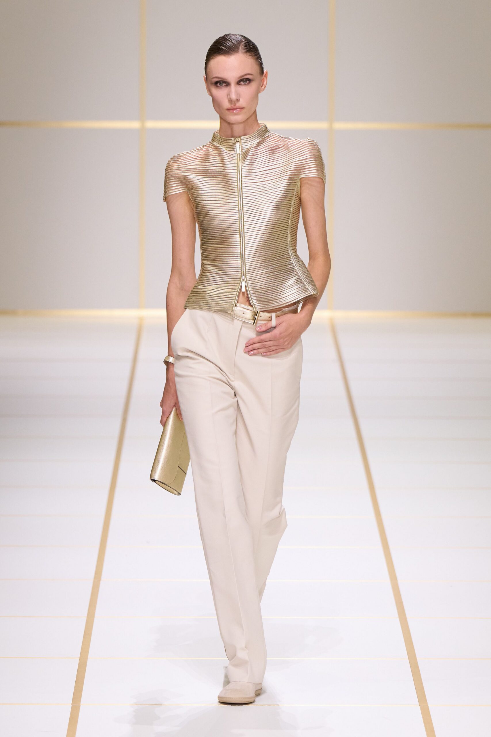Champagne ribbed top with a zip opening into a tiny hem split. Cream straight trousers with a pressed front crease. Narrow metallic belt, buckle set slightly off center. Plain gold cuff. Folded metallic envelope clutch. Pale square-toe shoes. For crossing a hotel lobby before the breakfast room opens.