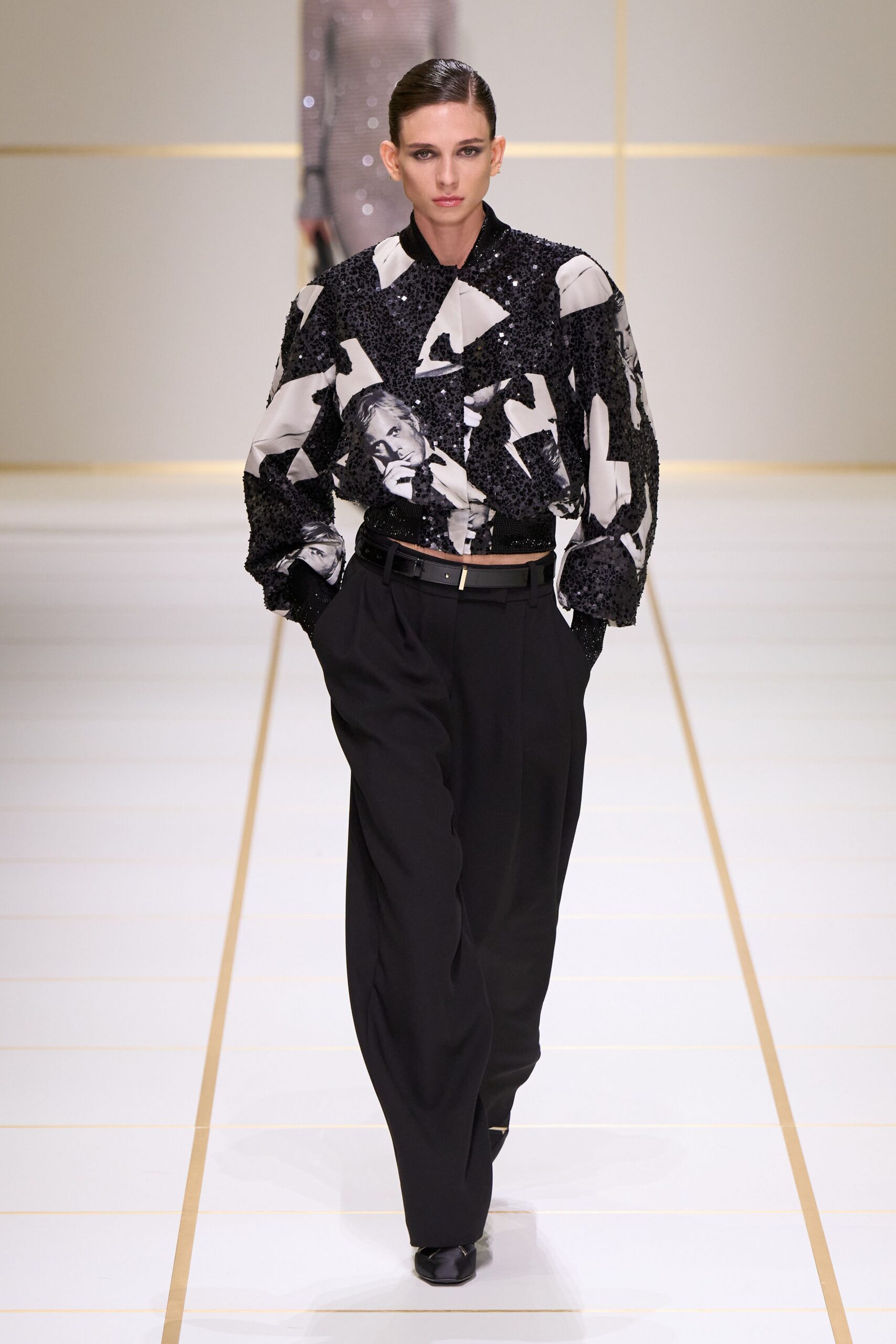Black sequined blouson spliced with monochrome portrait panels and a ribbed hem. Black pleated trousers, cut full through the leg. Slim black belt with a pale rectangular tip. Square-toe black shoes peeking under the break. Crossing a hotel atrium before the charity auction opens.