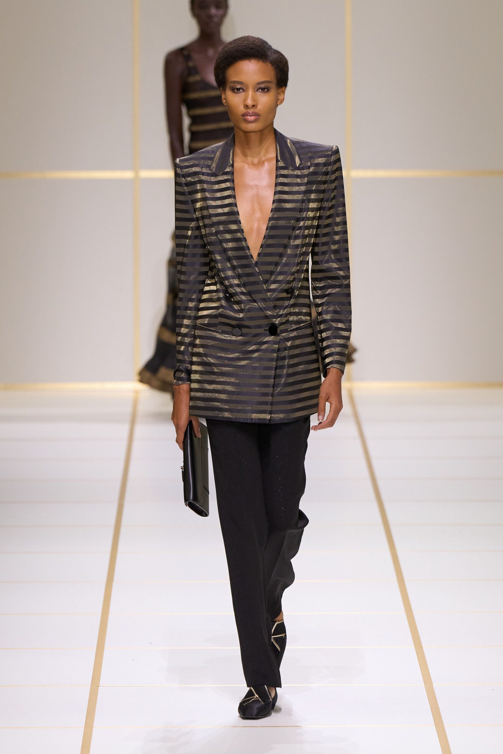 Black blazer shot with bronze horizontal stripes, cut to a plunging double-breasted wrap. Straight black trousers with a faint dry sparkle. Slim black clutch tucked flat against the thigh. Black pumps crossed with pale metallic straps. For arriving early to a hotel bar before the conference crowd.
