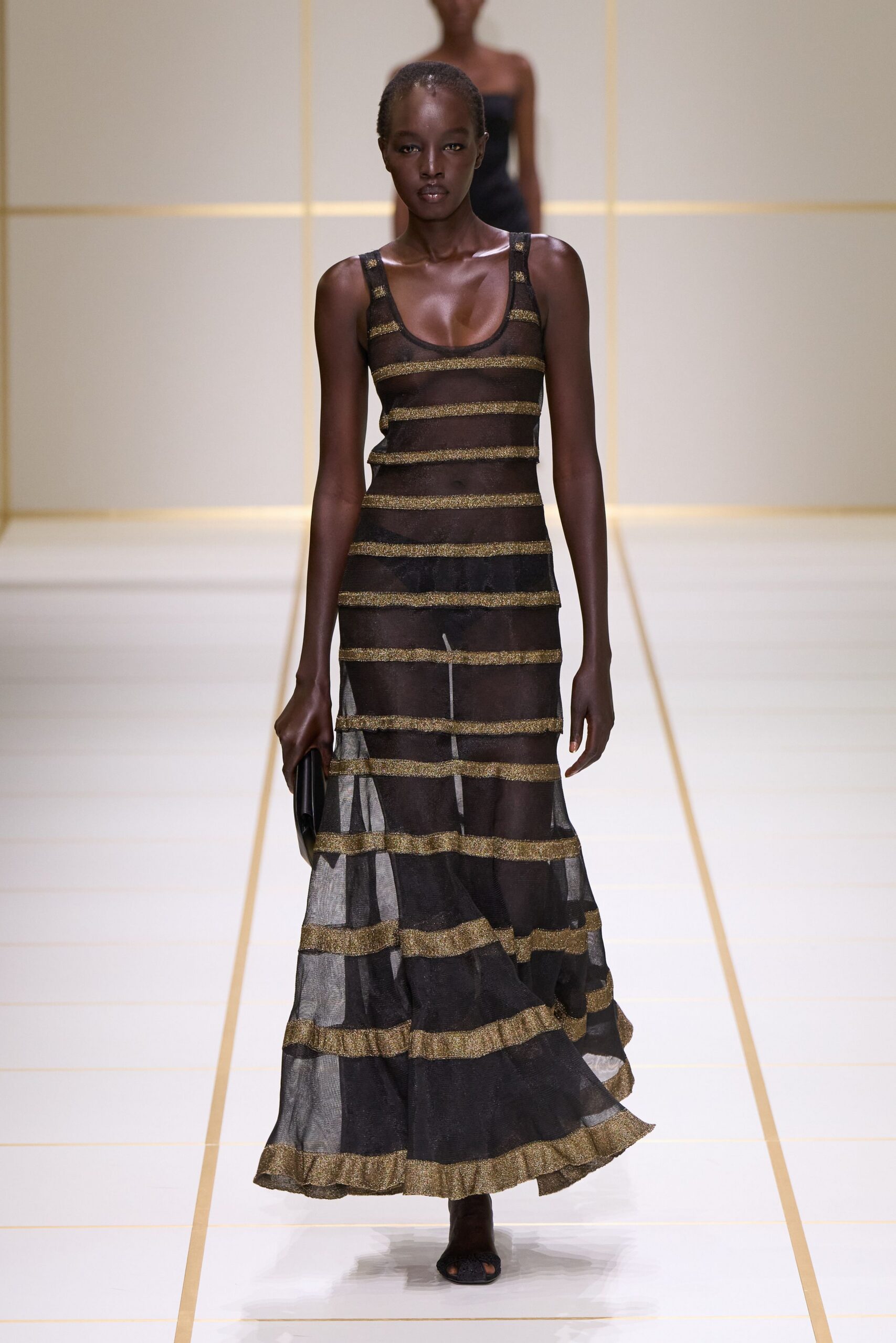 Sheer black mesh column dress banded in glittering gold stripes, hem kicked into a soft flounce. Flat black clutch tucked straight against the thigh. Black peep-toe heels, barely there under the skirt. Hotel terrace after midnight, the pool lights still making ladders on the tiles.