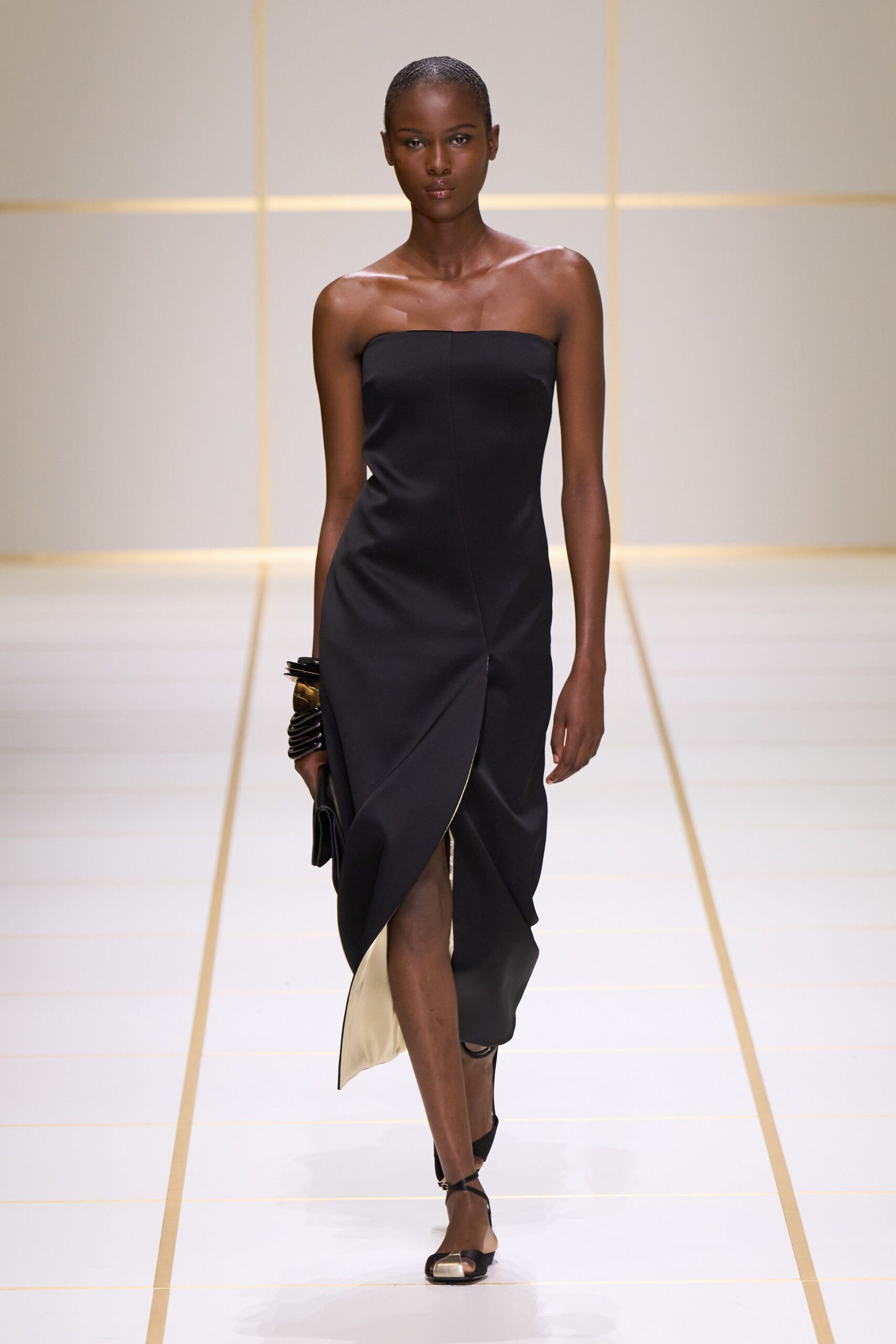 Black strapless column dress with a wrapped front slit and pale lining flash. Stacked dark resin bangles with one broad brushed-gold cuff. Flat black foldover clutch tucked under the wrist. Black ankle-strap sandals with a square silver toe cap. For arriving at a hotel bar before anyone else from dinner.