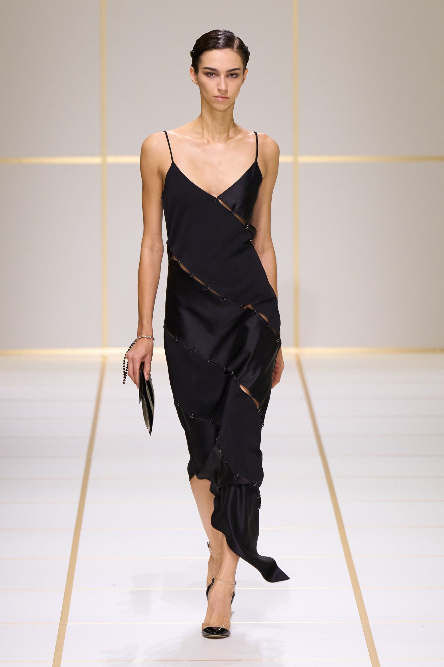 Black satin slip dress, spiral seams hooked open into narrow slashes. Flat black clutch with a beaded wrist loop. Black patent slingbacks with pointed toes and clear sides. Crossing a hotel atrium after midnight with the room key in hand.