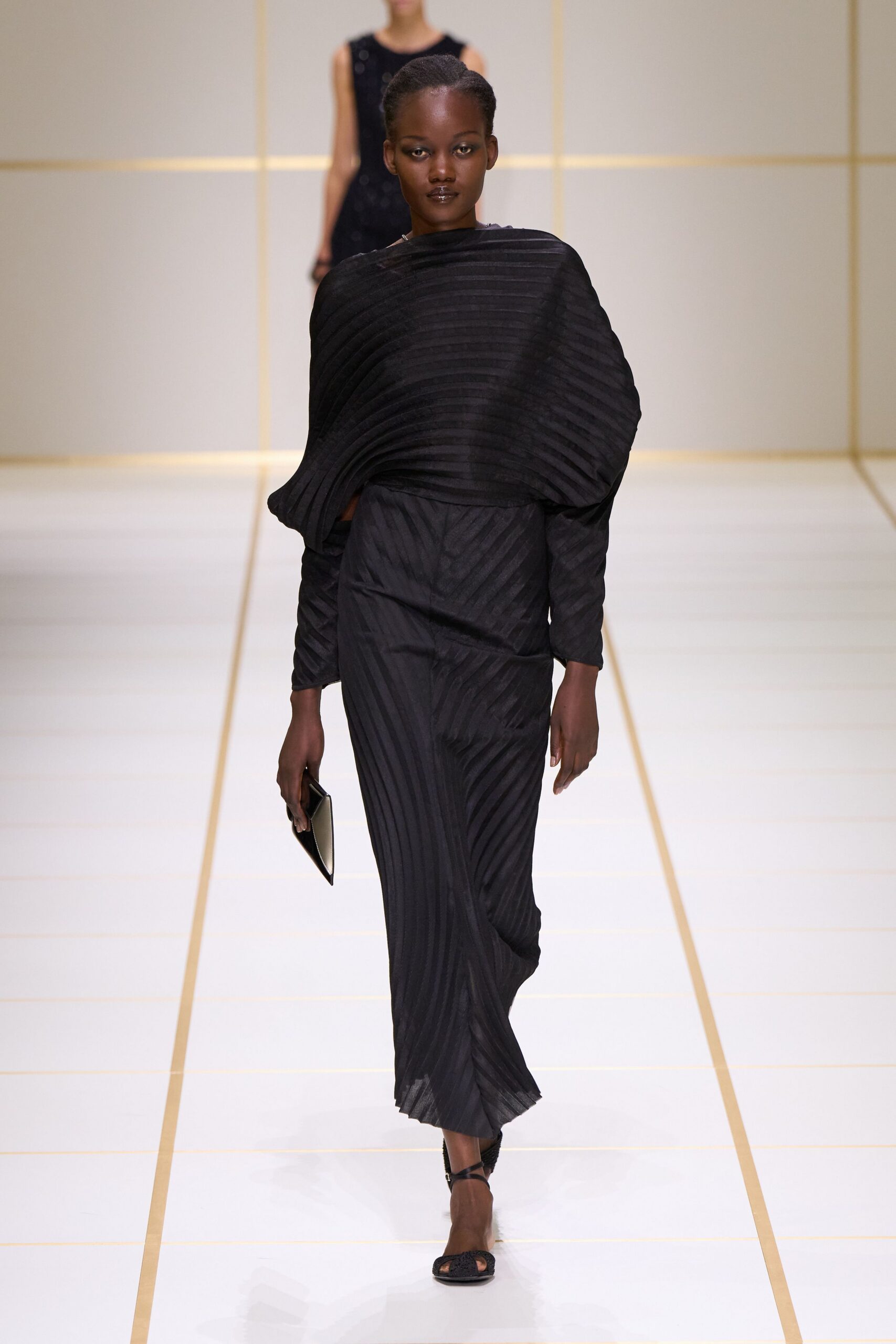 Black plisse top wrapped into a sculpted one-shoulder cocoon. Matching bias-striped plisse skirt, hem opening into narrow knife pleats. Flat black envelope clutch. Black ankle-strap sandals. Crossing a silent hotel atrium before the conference crowd comes down.