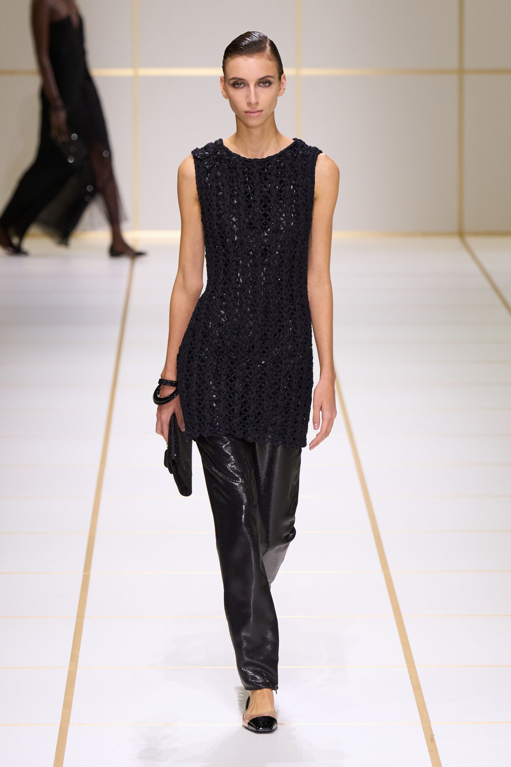 Black sleeveless tunic in open braided lattice, narrowing sharply at the waist. Black glossy trousers with a faint reptile sheen down the leg. Stacked black lacquer bangles. Soft black beaded clutch tucked low in one hand. Black cap-toe pumps with clear vinyl sides. Crossing a hotel atrium before the string quartet starts.