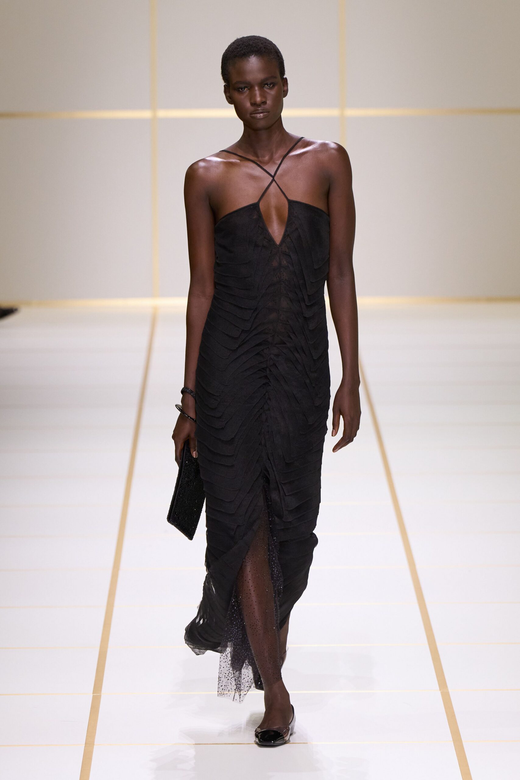 Black column dress with crossed spaghetti straps and chevron tiers of ruched chiffon. Sheer dotted tulle panel surfacing through the front slit. Two slim black bangles. Beaded black clutch tucked flat in one hand. Black patent cap-toe flats. Crossing a hotel atrium before the strings begin tuning.