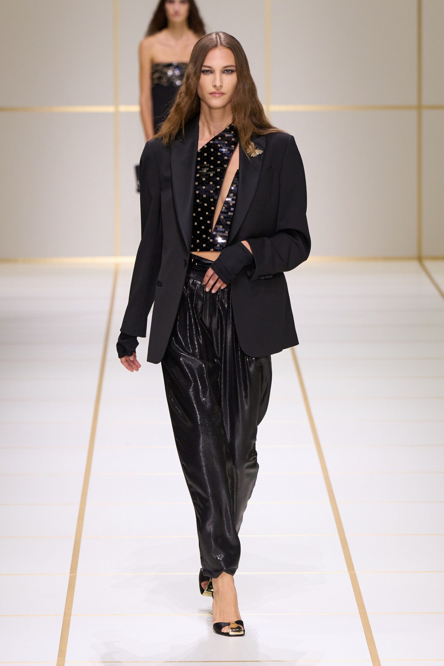 Black blazer with satin lapels and a gold insect brooch. One-shoulder sequined top, cut to a deep bare V. Loose black trousers in wet-look cloth, pooling into soft creases. Black peep-toe mules with square gold-capped fronts. Walking into a hotel bar after midnight with one earring missing.