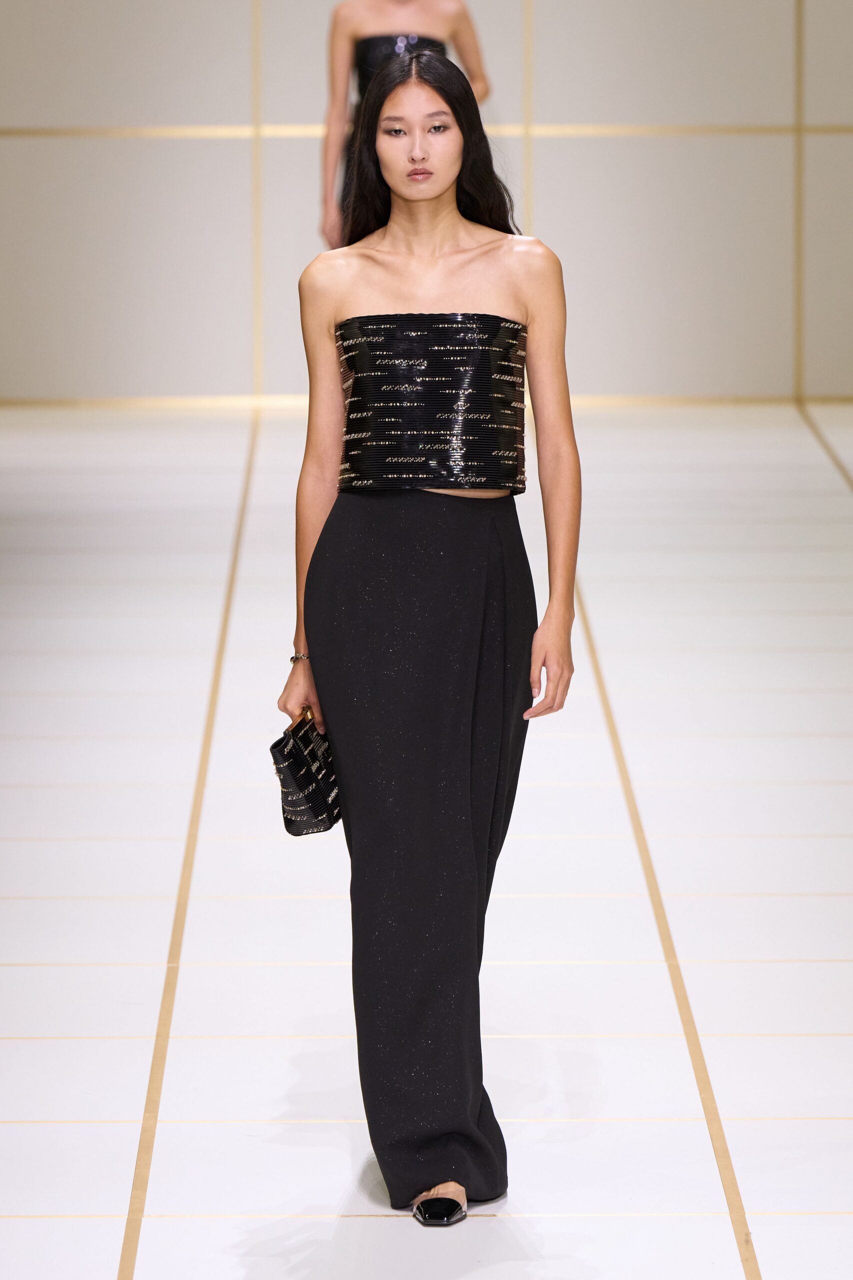 Black sequined tube top banded in narrow horizontal rows. Slim silver bracelet at one wrist. Black column skirt with a wrapped panel and faint glitter. Matching beaded clutch with a small wooden handle. Black patent square-toe flats. Hotel ballroom carpet just before the awards begin.