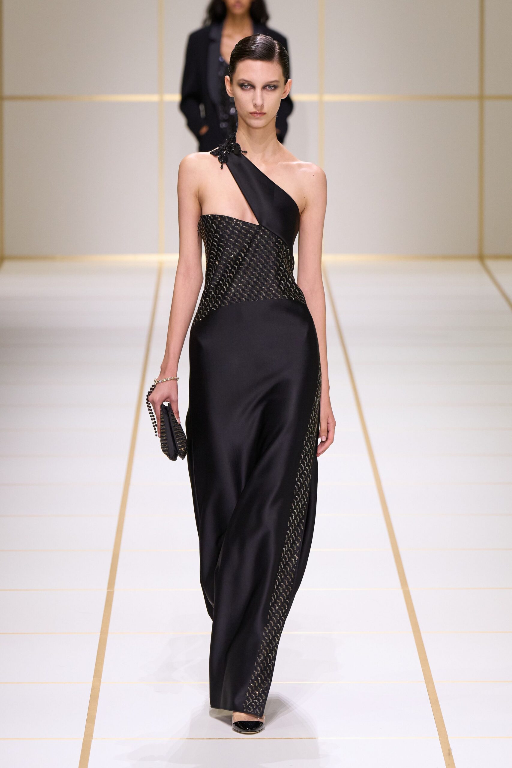 Black satin one-shoulder column gown, lattice bodice curving into a bare side. Beaded black flower pinned at the single strap. Pleated black clutch with a looped bead handle. Black patent pumps peeking from the hem. For arriving at a hotel bar after the wedding crowd has thinned.
