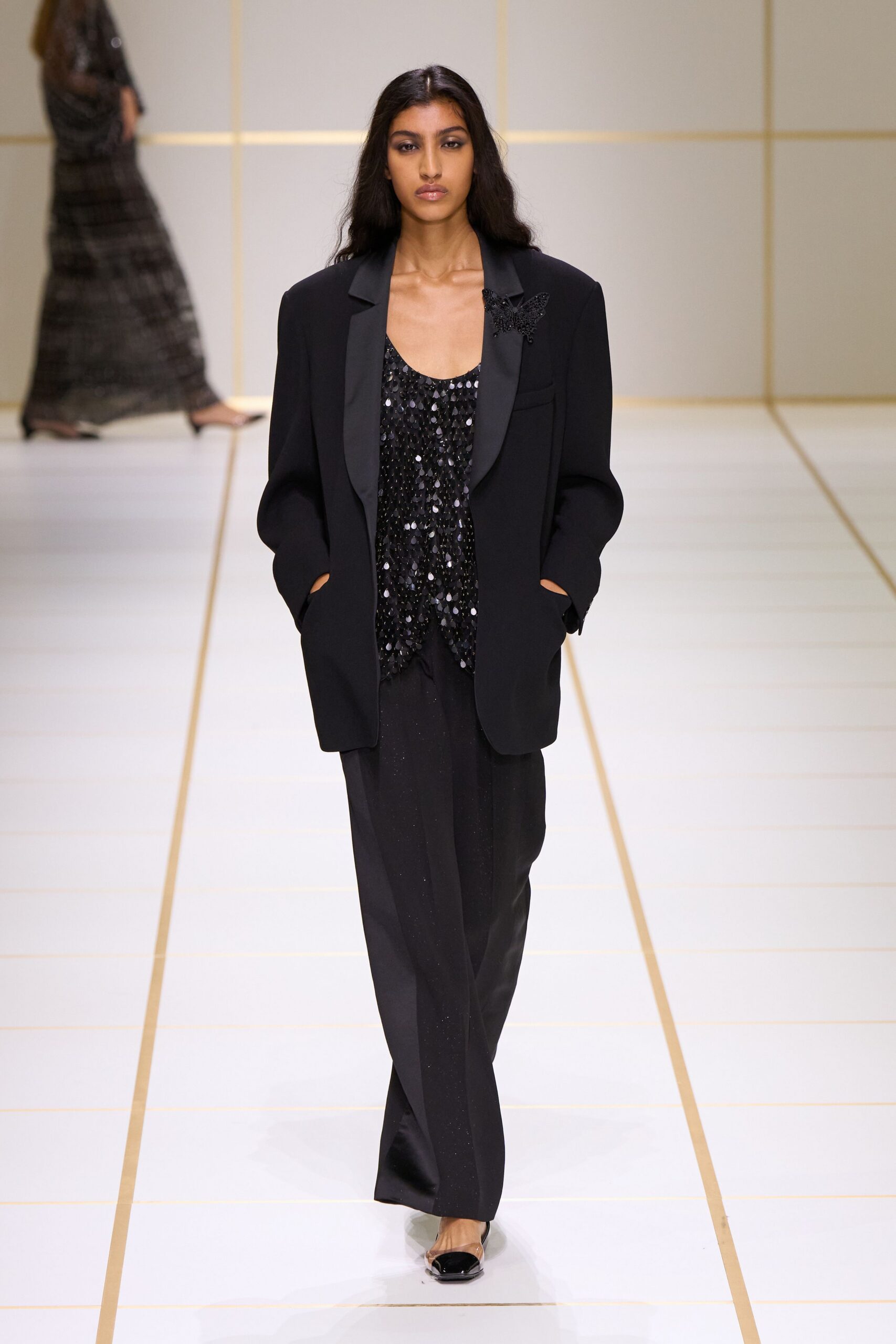 Oversized black blazer with satin shawl lapels and a beaded butterfly brooch. Black scoop-neck shell densely covered in glossy paillettes. Loose black trousers with a faint glitter through the crease. Black cap-toe pumps. Walking into a hotel ballroom after the chairs have been stacked.
