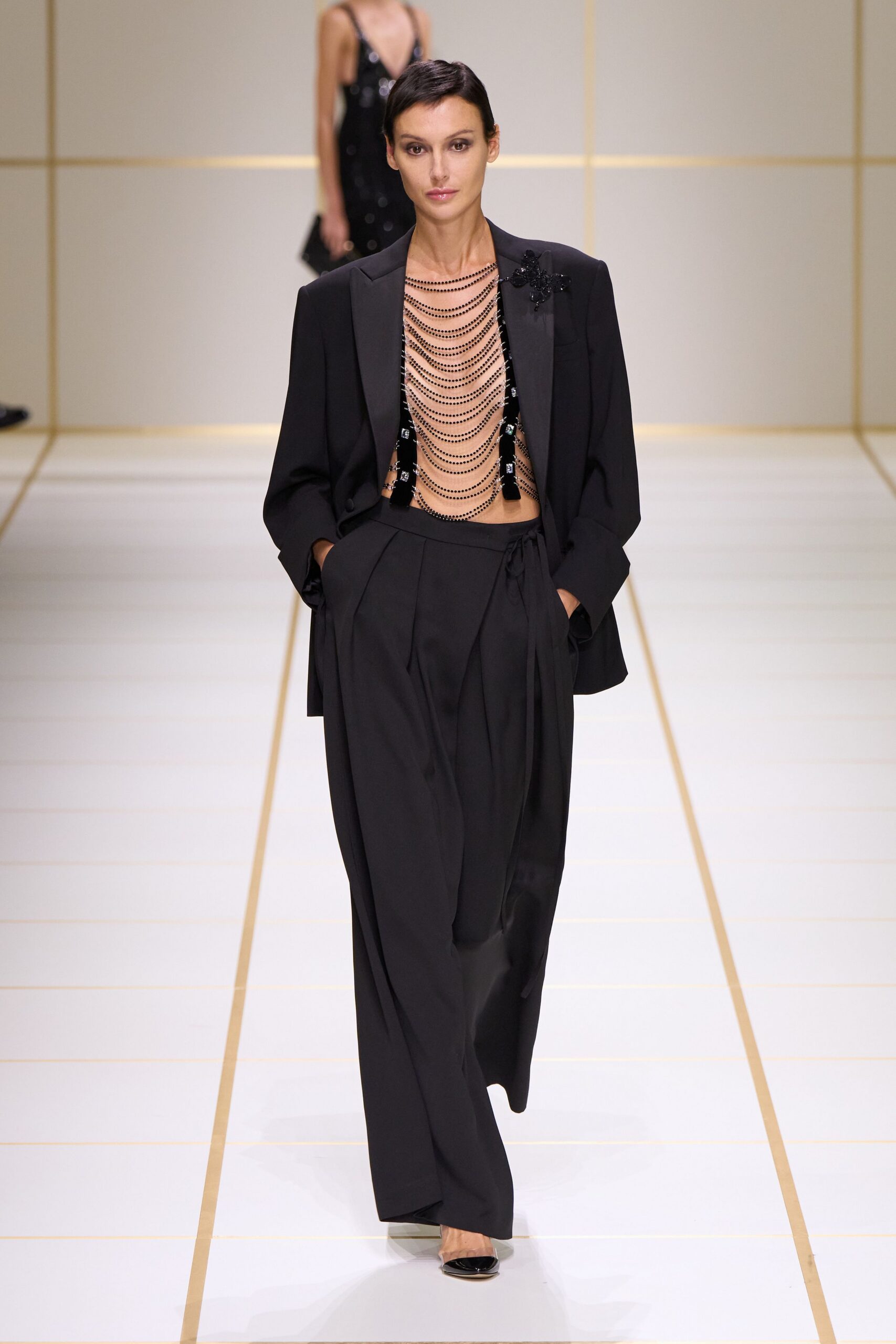 Black tuxedo blazer with satin lapels and a beaded flower on one shoulder. Black beaded chains draped in deep scallops across the bare chest. Fluid black wrap-front trousers with a long sash falling off-center. Black patent pointed pumps. For slipping out of the hotel bar before anyone reaches the bill.