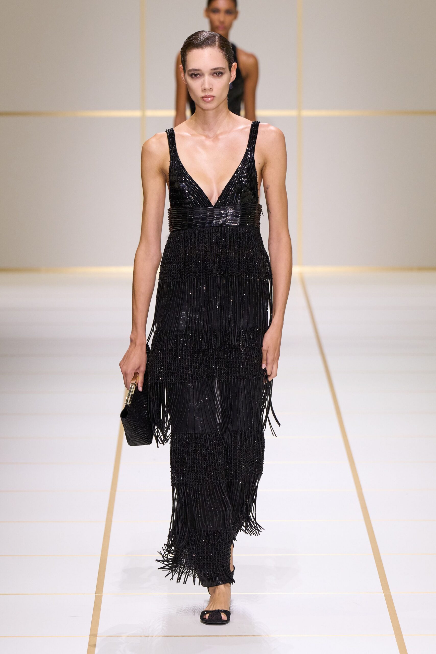 Black beaded column gown with a plunging V and long fringe tiers. Small black clutch with a gold-toned frame clasp. Black peep-toe mules, the front strap dusted with beads. Crossing a hotel atrium after midnight, hem still shivering from the stairs.