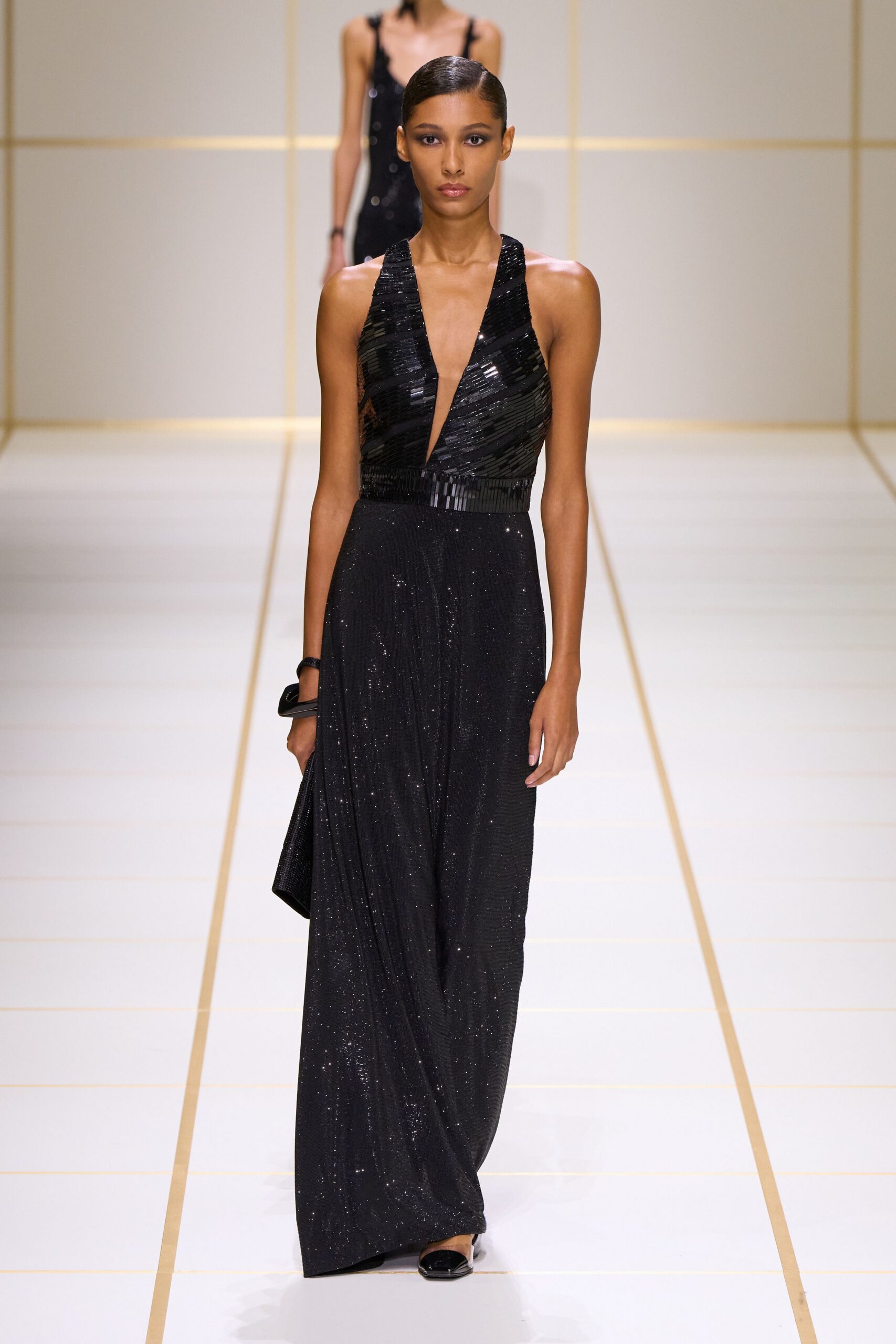 Black evening gown with a plunging halter bodice in horizontal bugle beading. Slim black clutch tucked under one hand. Square-toe black mules with a silver metal heel cap. Hotel ballroom carpet after midnight, band still packing up.