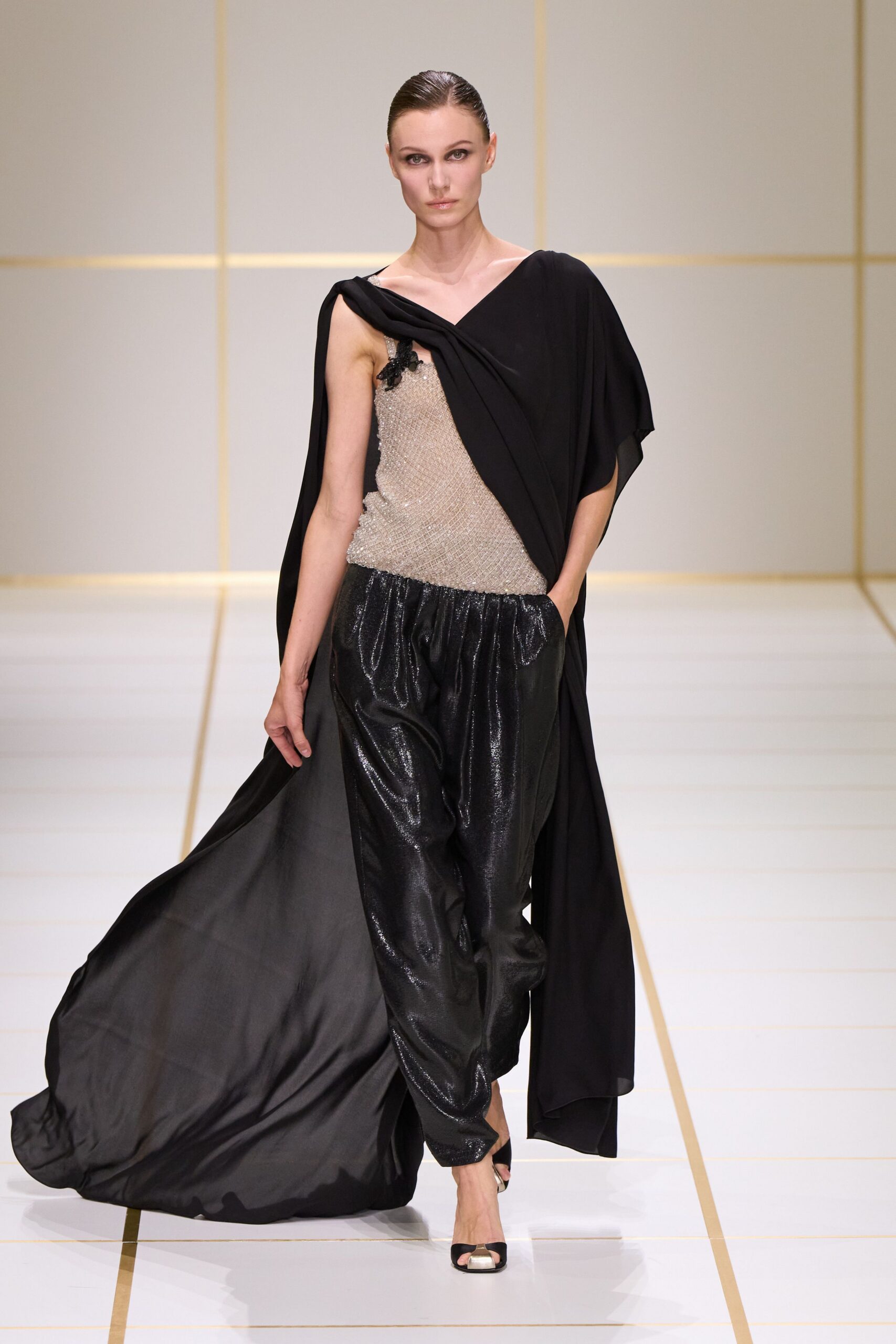 Black crepe drape crossing one shoulder and trailing to the floor. Silver beaded mesh camisole with a black floral applique at the strap. Black liquid-shine harem trousers, gathered softly into the waist. Peep-toe mules with a pale metallic cap. For the hotel ballroom after the auction tables have been cleared.