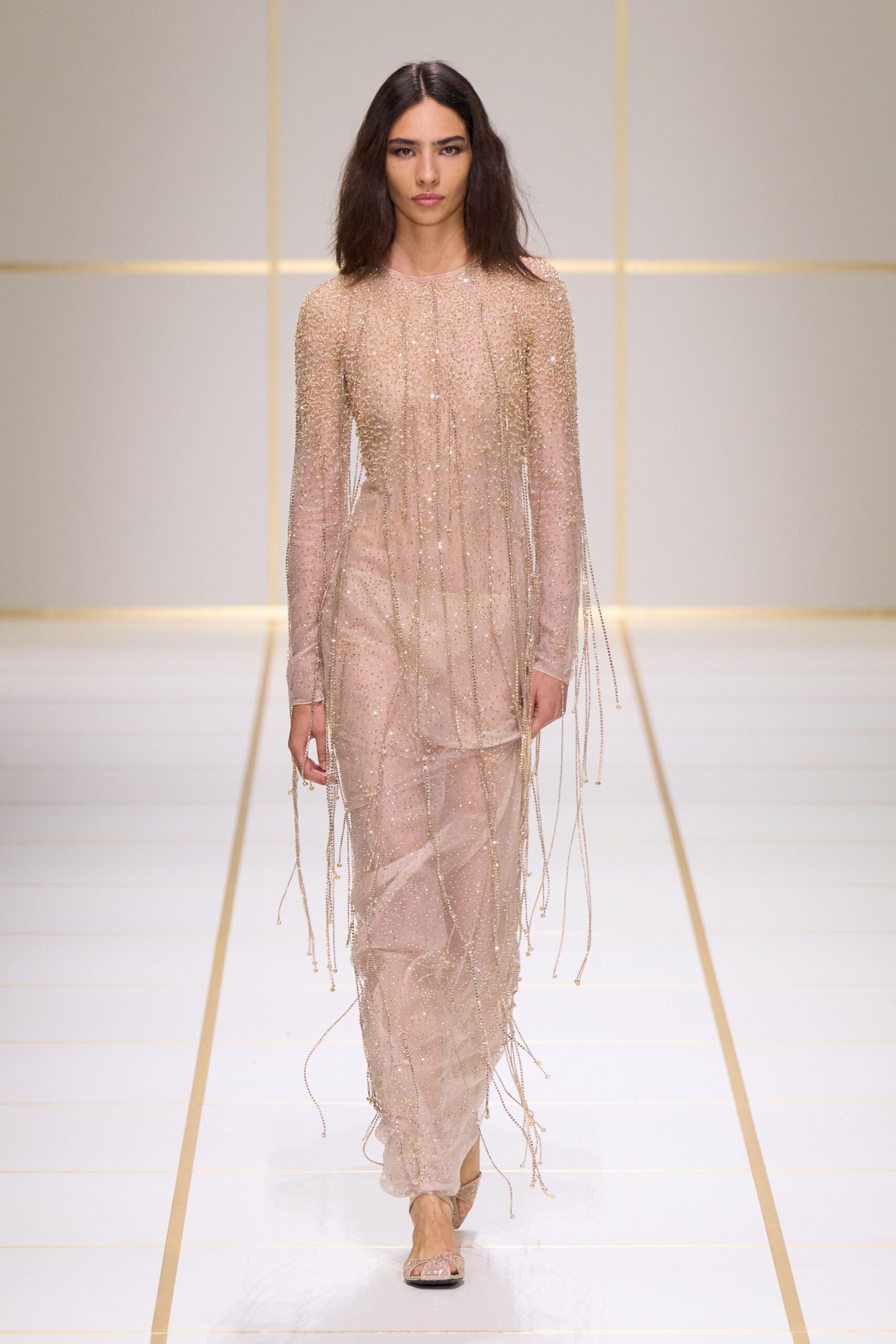 Blush sheer gown dusted in crystals, with bead-chain fringe falling past the hands. Nude satin sandals with glittering mesh over the toes. Crossing a hotel atrium just after the strings have packed up.