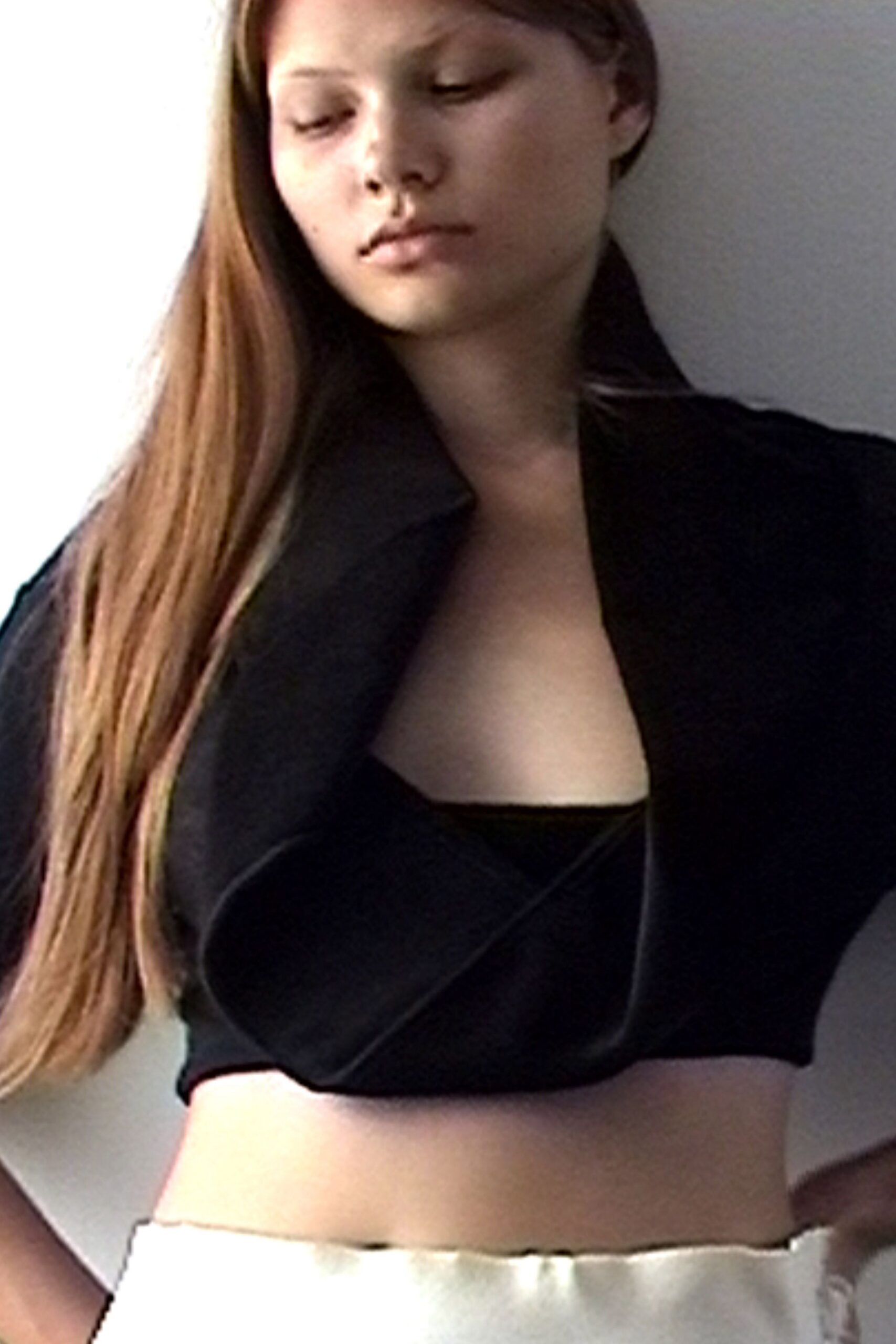 Black cropped jacket with broad rolled lapels falling open at the chest. Black bandeau top, cut straight across. Ivory satin skirt with a faintly rippled waistband. Afternoon apartment light, halfway dressed before answering the door.