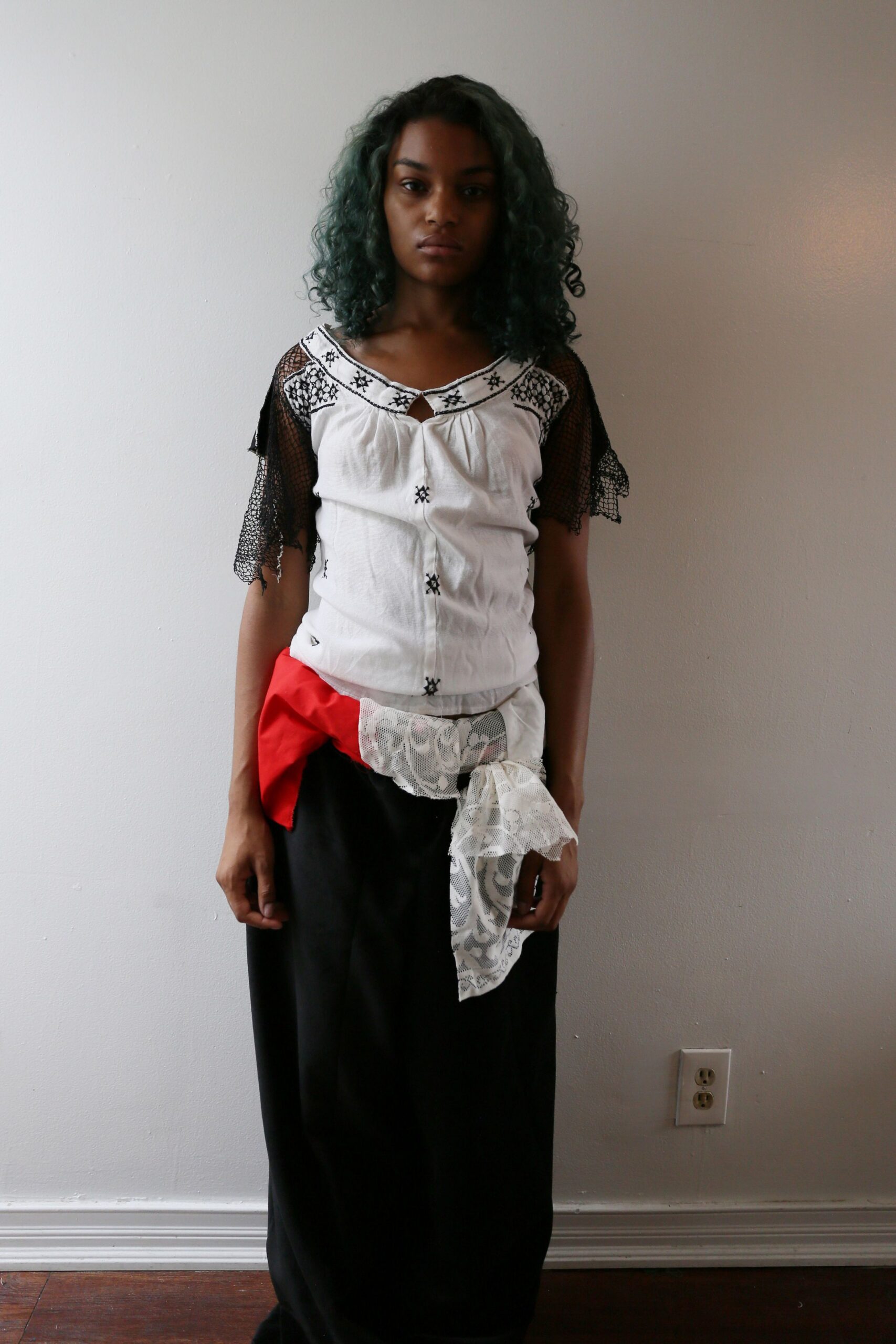 White peasant blouse with black embroidery and loose net flutter sleeves. Red fabric knot spliced with white floral lace at the hip. Long black column skirt, hanging straight to the floor. Walking home from a street festival after the folding chairs are stacked.
