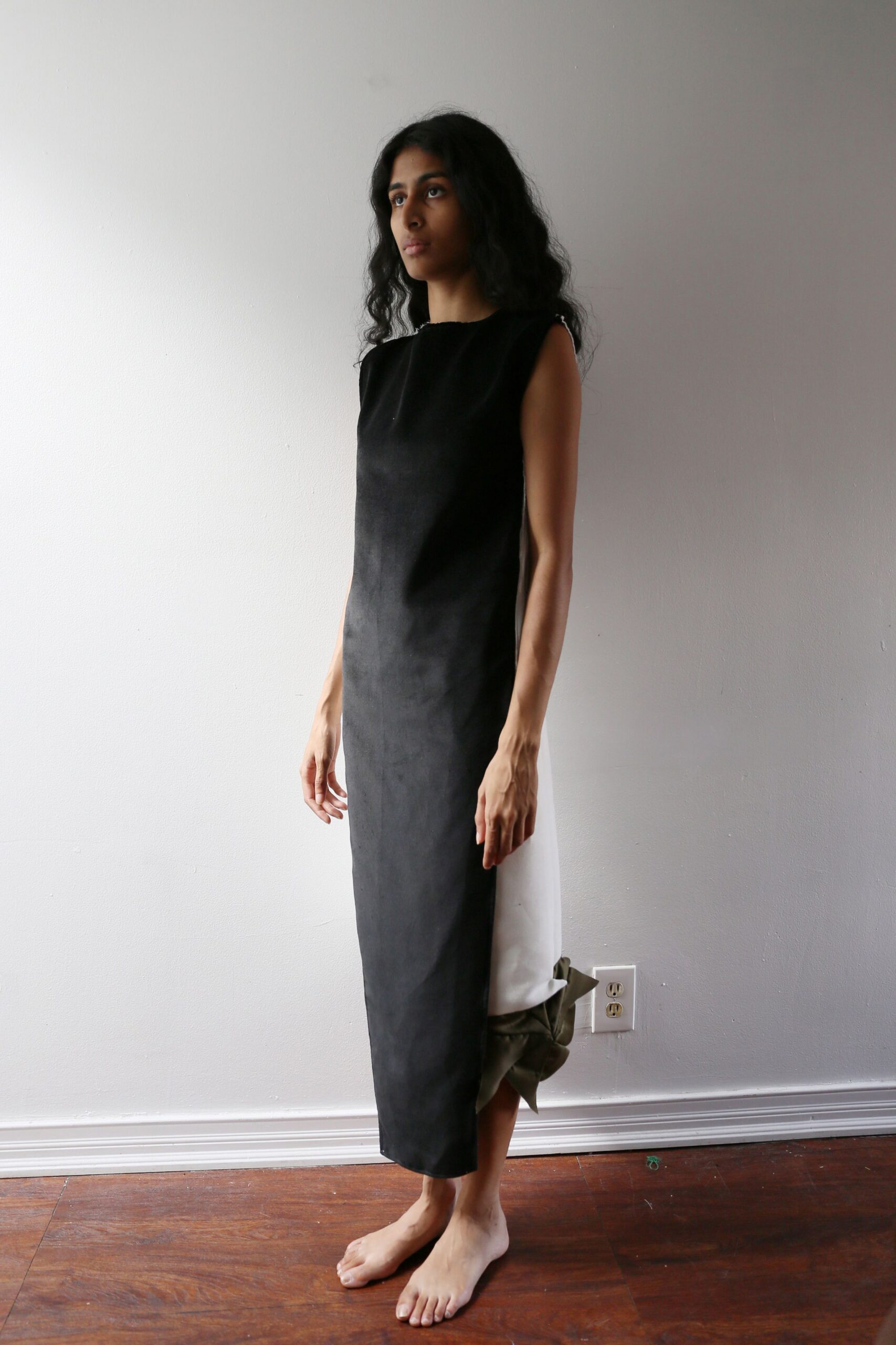 Black column dress in matte sueded cloth, white side panel slipping loose at the calf. Olive fabric bundle caught at the back like a collapsed bow. Afternoon apartment light, deciding whether to answer the downstairs buzzer.