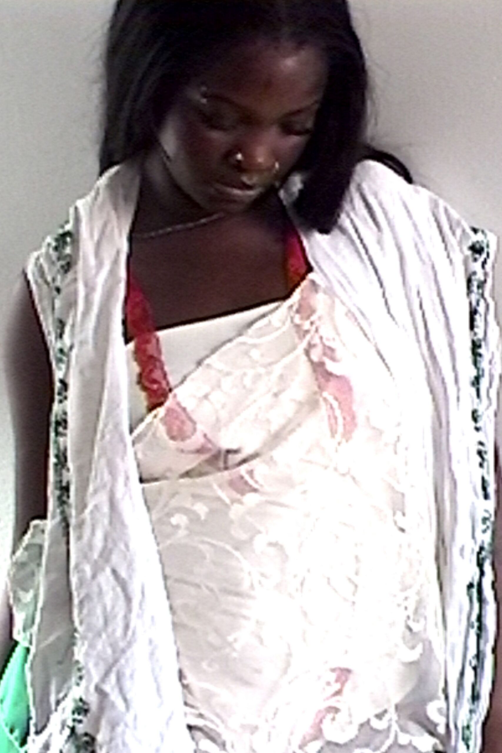 Ivory satin slip dress, pink flocked scrolls crossing the bust. White crinkled shawl with narrow green floral bands at the edges. Red lace bra peeking above the slanted neckline. Fine silver chain at the collarbone. Twin silver nose hoops. Afternoon heat in a borrowed room, curtain half pinned shut.
