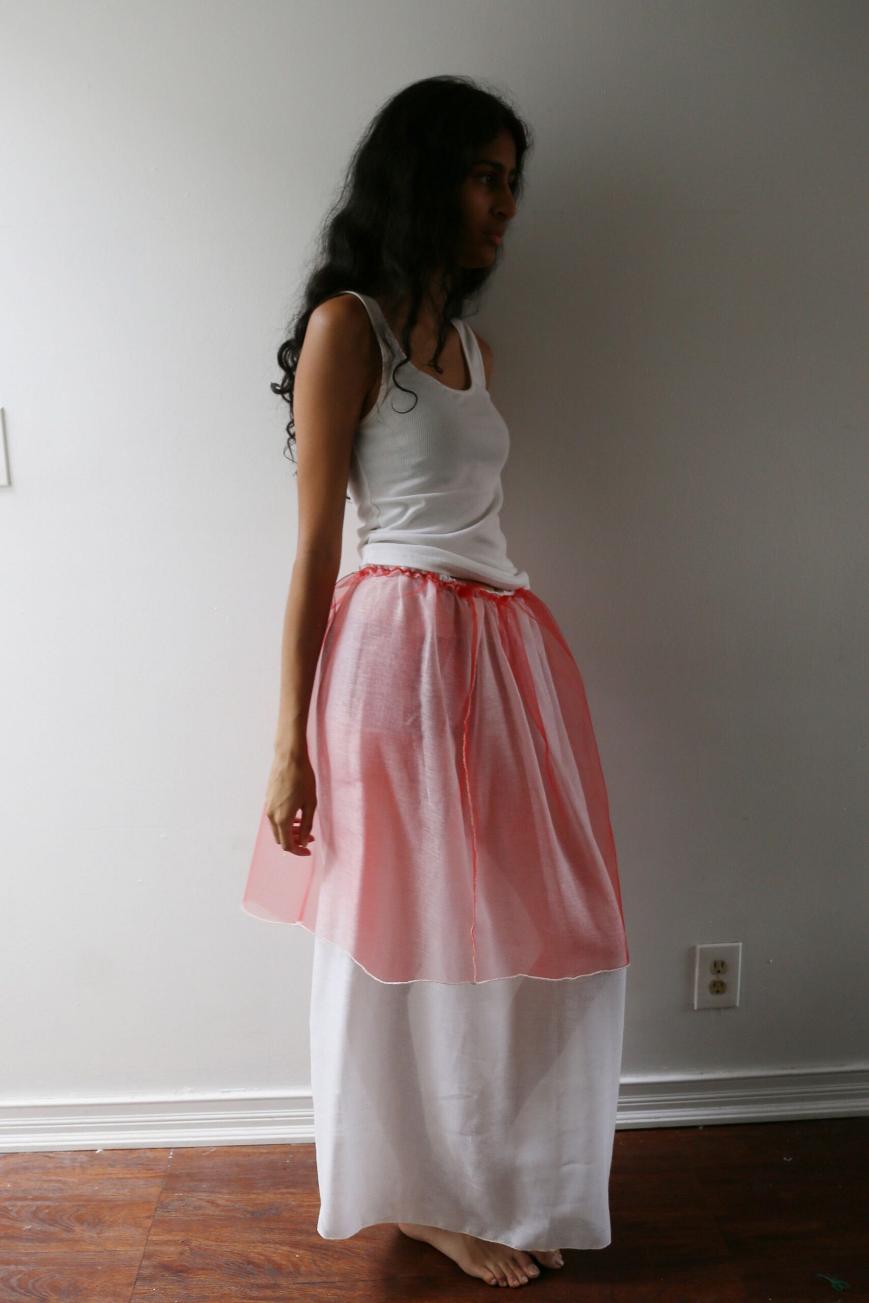 White ribbed tank, scoop neck sitting close to the collarbone. Sheer coral organza overskirt, gathered on a narrow drawstring waist. Long white skirt in airy plainweave, falling straight to bare feet. Morning light in an empty rental before the boxes arrive.