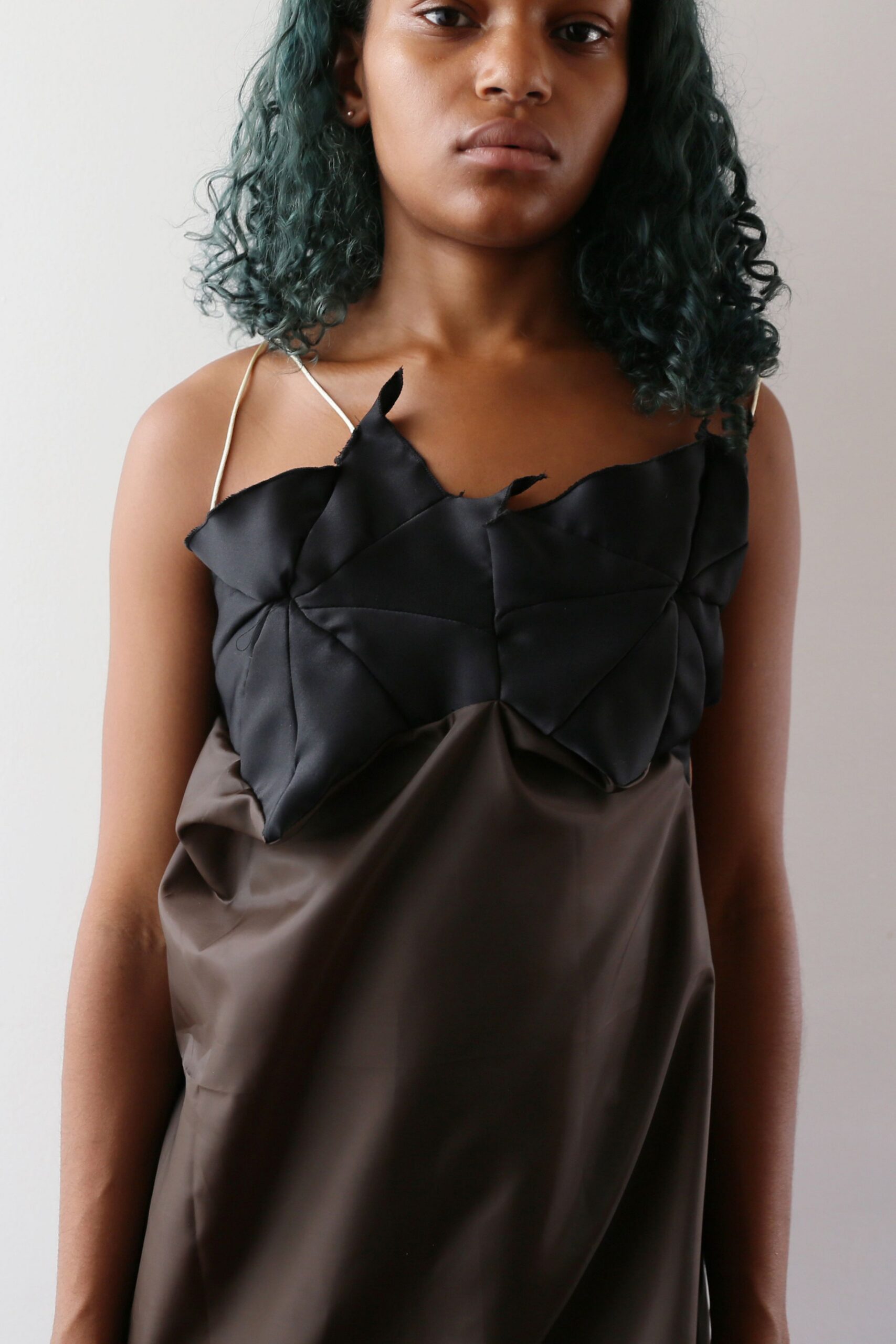 Brown satin slip dress with thin cream straps and a collapsed drape. Black folded satin panels across the chest, raw points lifting at the edge. Tiny silver studs. Morning in a borrowed apartment, dress still creased from the suitcase.
