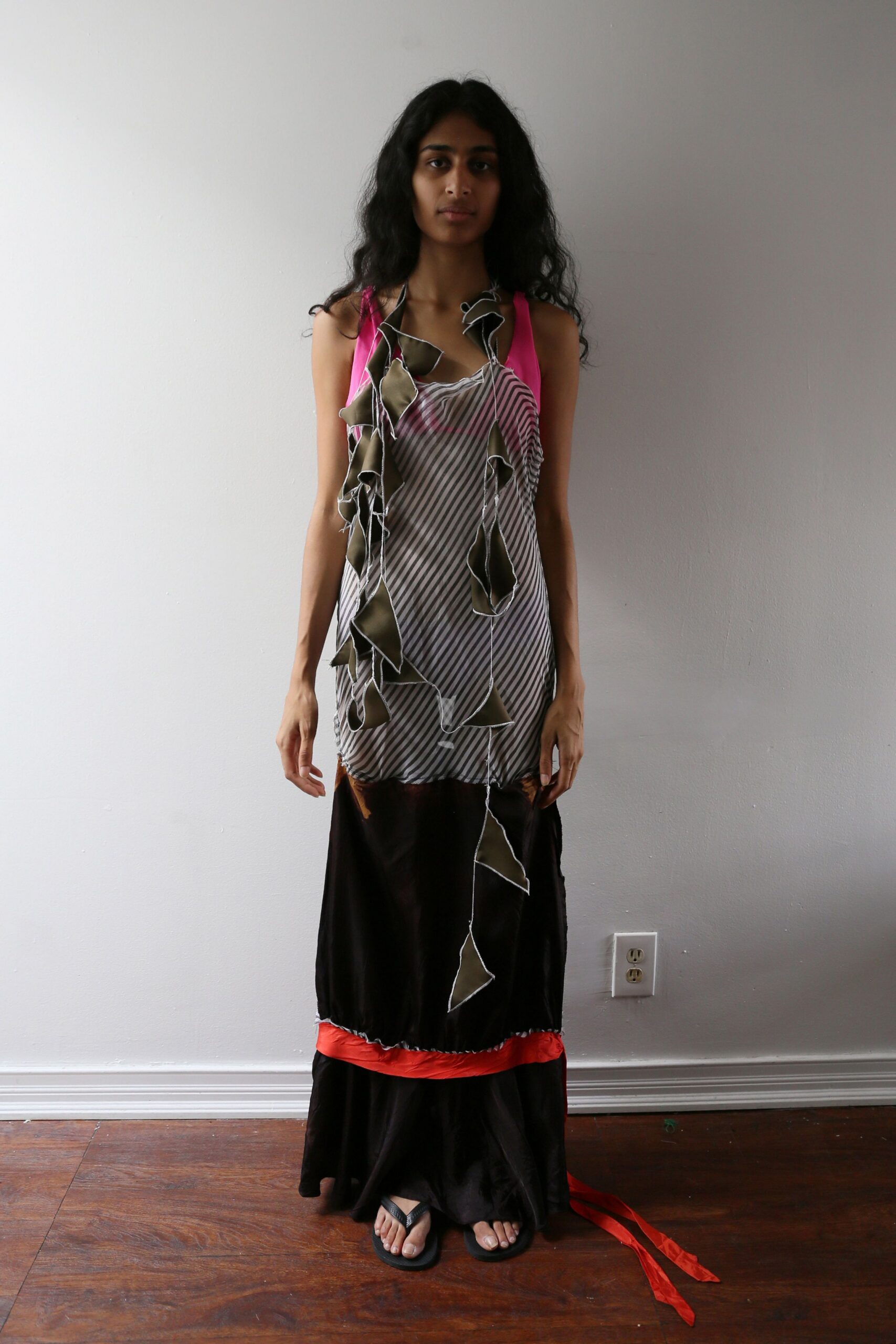 Bias-striped slip dress with raw overlocked appliques hanging in loose ribbons. Black satin panel peeking below, finished with a tomato-red ruffle. Black flip-flops, flat and plain. For crossing a bare apartment with packing tape still on the floor.