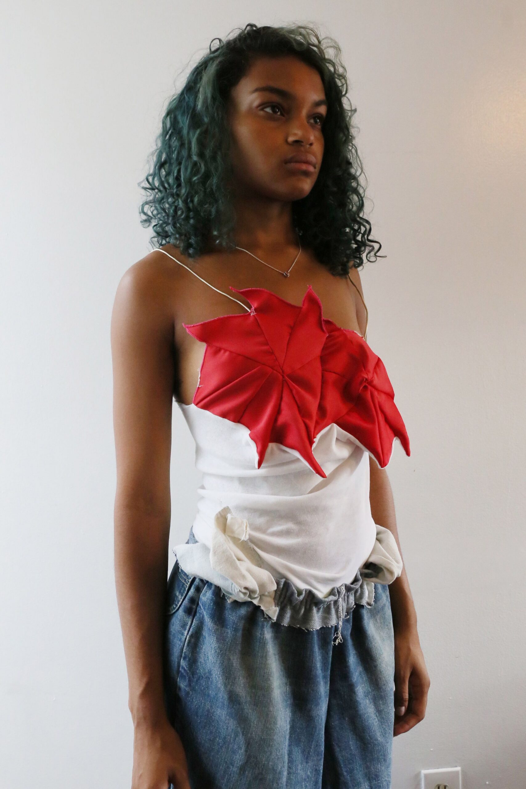 Ivory camisole with wire-thin straps and a red origami flower bust. Faded indigo denim trousers with a ruffled grey drawstring peeking up. Fine silver chain with a tiny dark heart pendant. Morning in a borrowed apartment, packing before anyone else wakes.