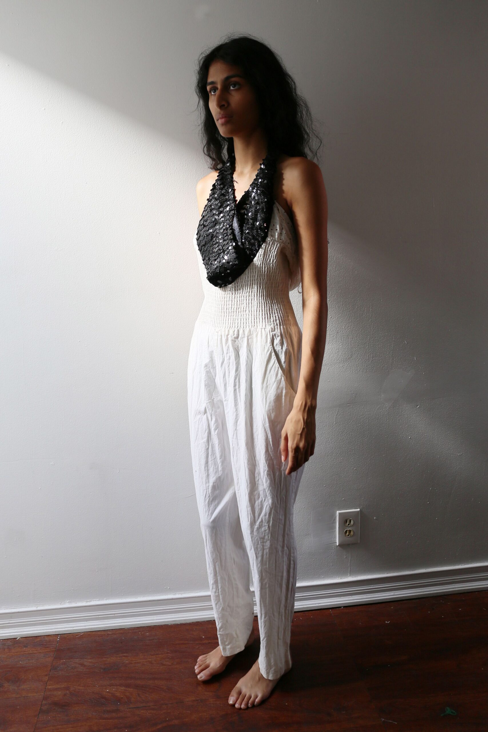 Black sequin scarf looped low, one end hanging longer. White cotton jumpsuit with a shirred bodice and crumpled straight legs. Morning light in a borrowed apartment before anyone finds the coffee.