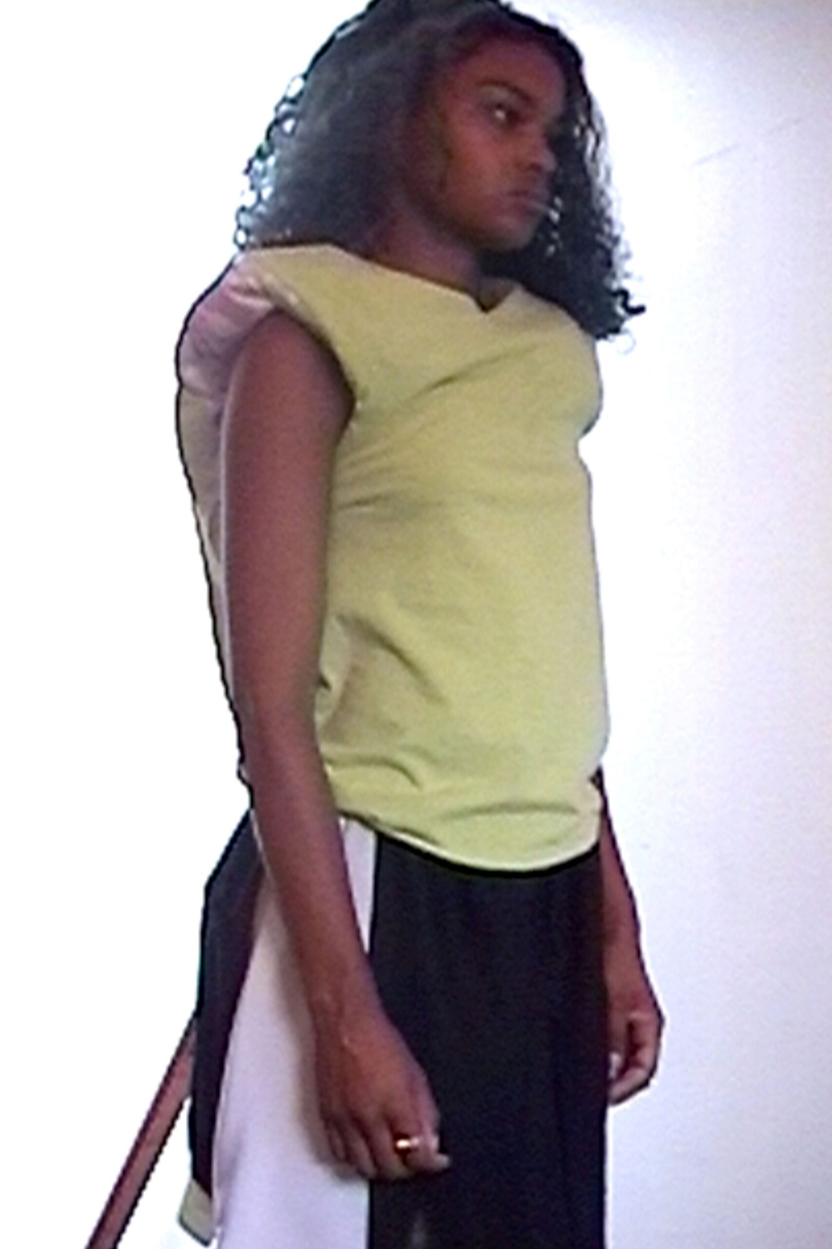 Pale chartreuse jersey top with padded cap shoulders and a small notch neckline. Black trousers sliced with an ivory side panel and one hanging strap. Plain gold band on one finger. Waiting by the freight elevator after dropping off film at a lab.
